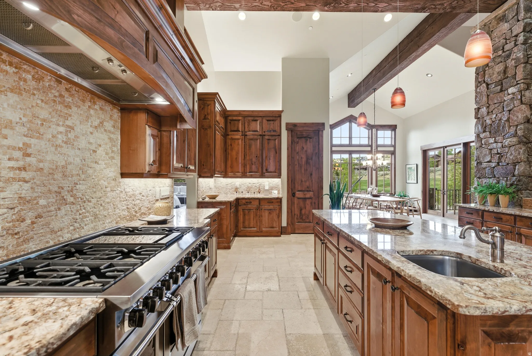 Spacious kitchen featuring an expansive island with an undermount sink, a professional-grade stainless steel range with a brick-finish backsplash, wood cabinetry, and vaulted ceilings with exposed wood beams