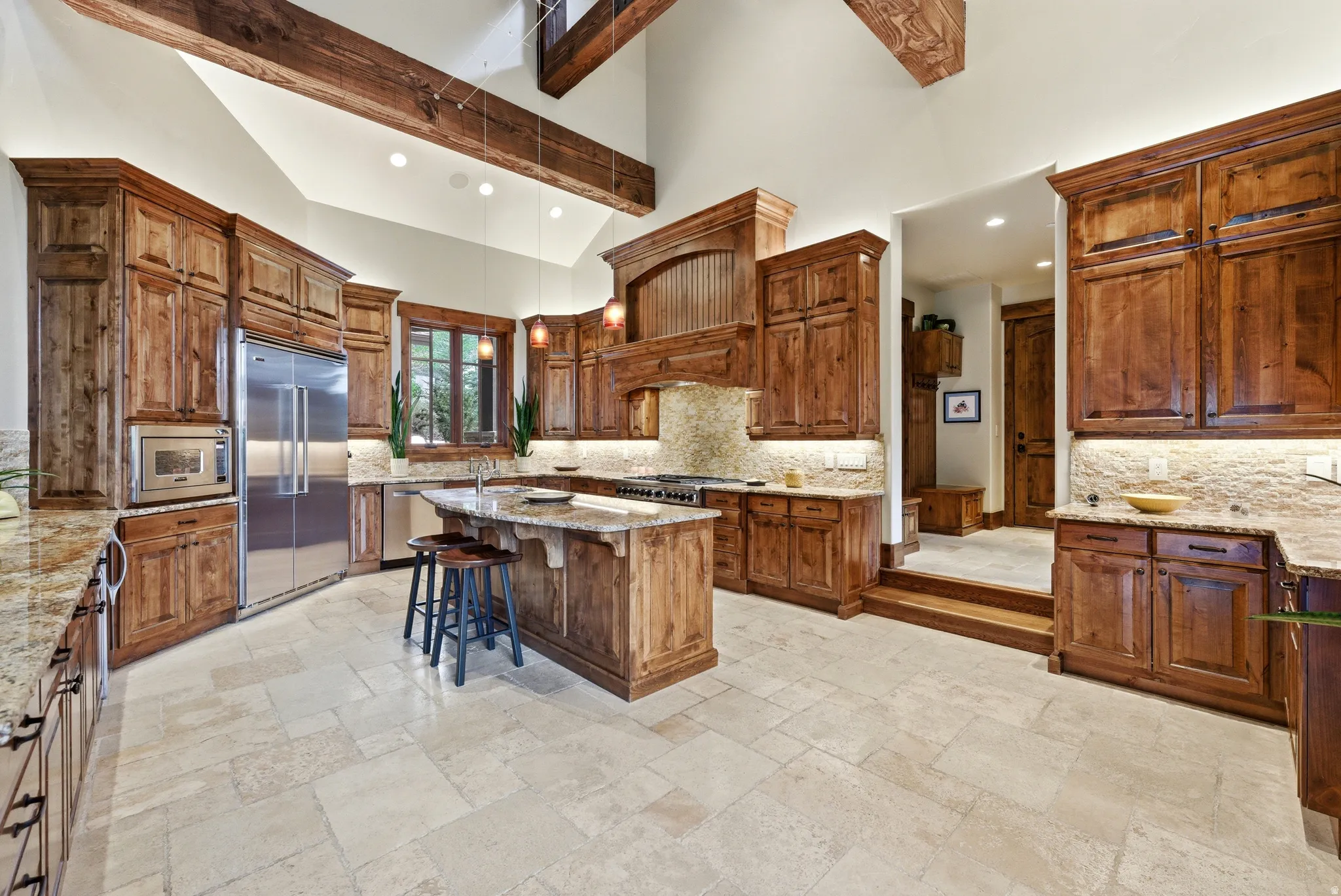 Spacious kitchen featuring custom wood cabinetry, stone countertops, integrated stainless steel appliances, a center island with seating, and vaulted ceilings with exposed wood beams