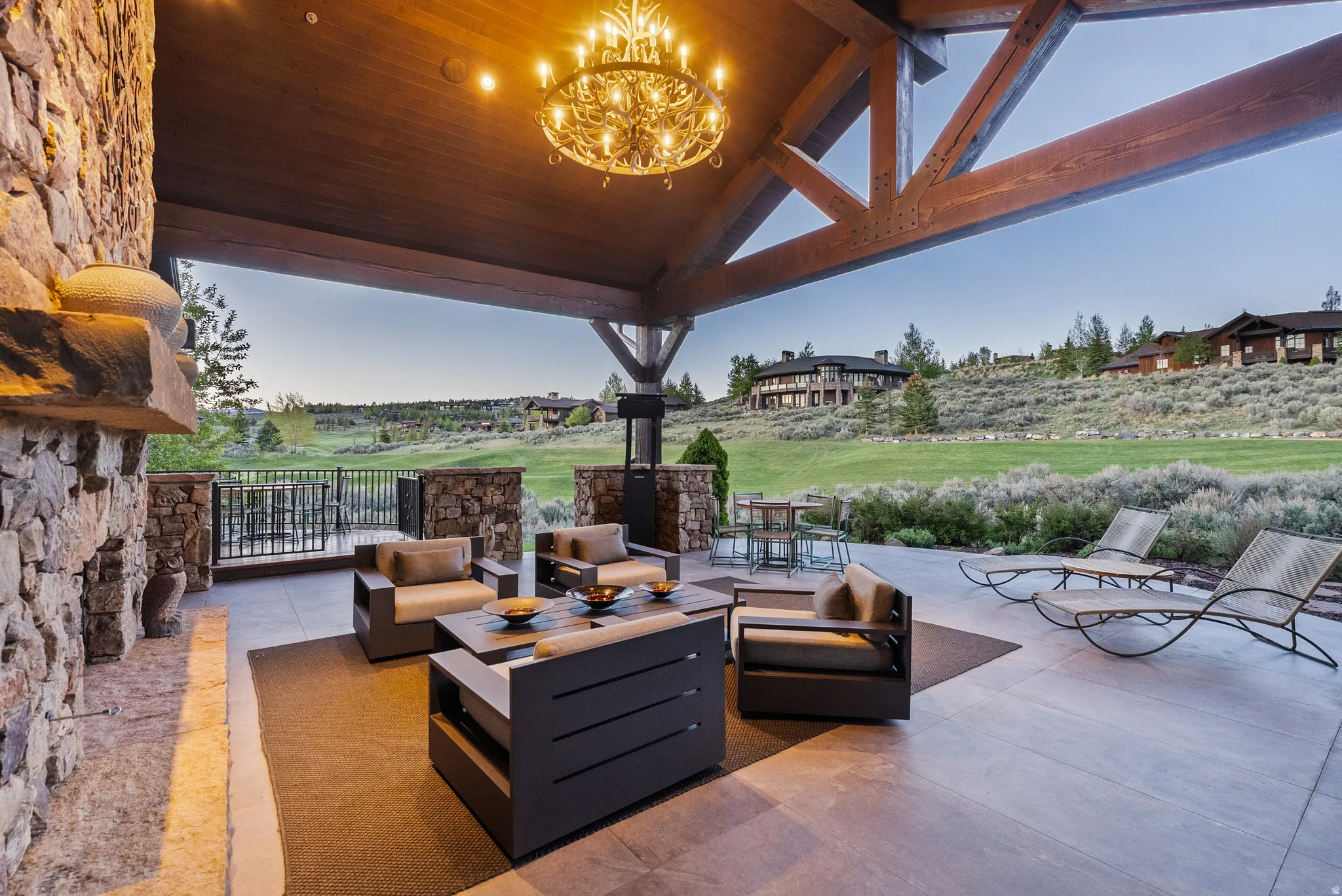 Expansive outdoor covered patio featuring a stone fireplace, exposed timber beams, a decorative chandelier, and a concrete patio surface