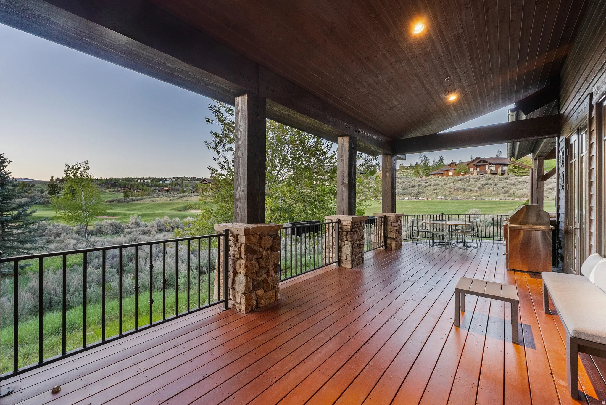 Expansive covered deck featuring rich wood-finish flooring, sturdy stone-wrapped columns, a built-in stainless steel grill, and a dark metal railing