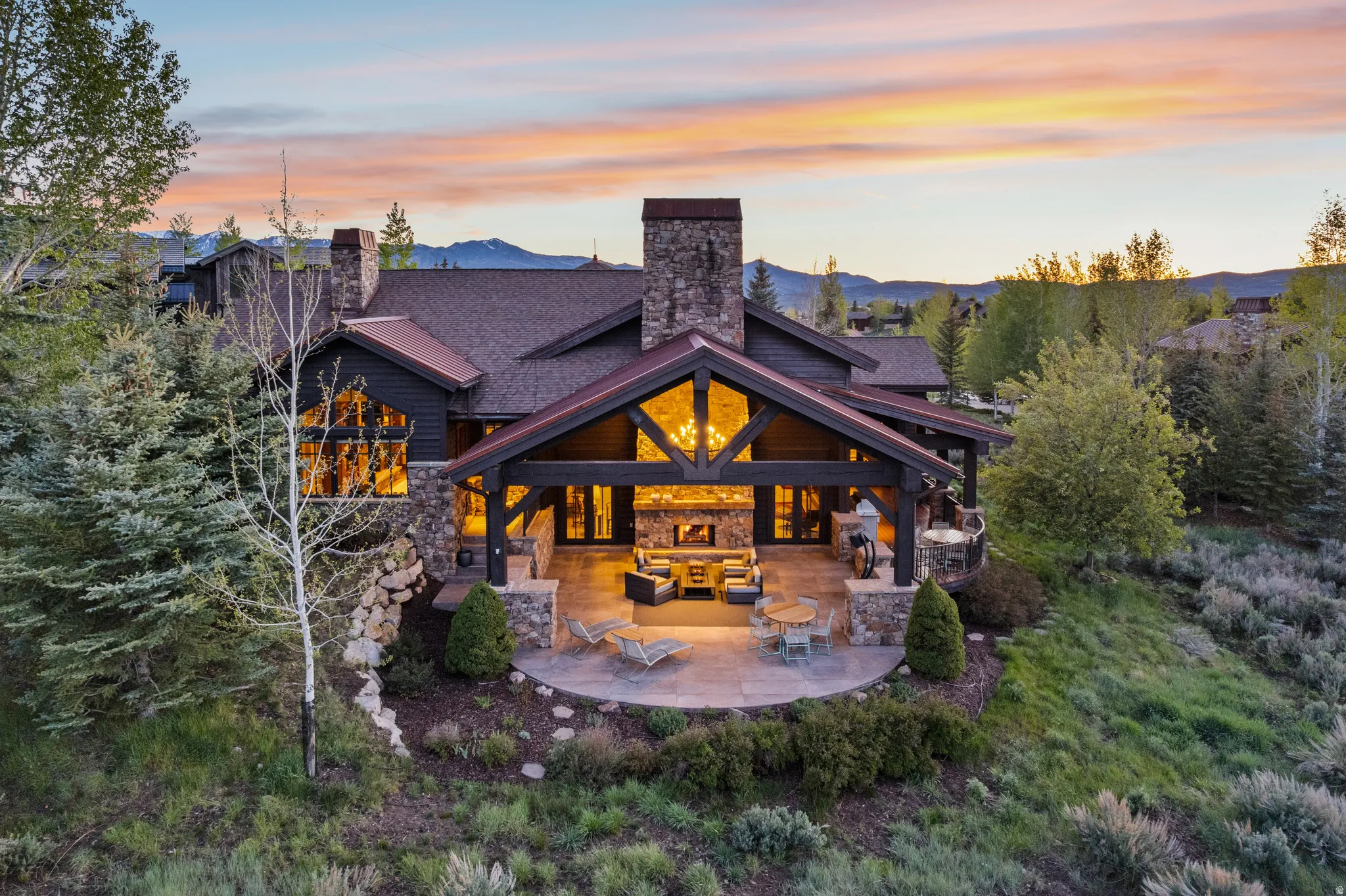 Stone and timber frame exterior featuring a large covered patio with an outdoor fireplace, a stone retaining wall, and mature landscaping