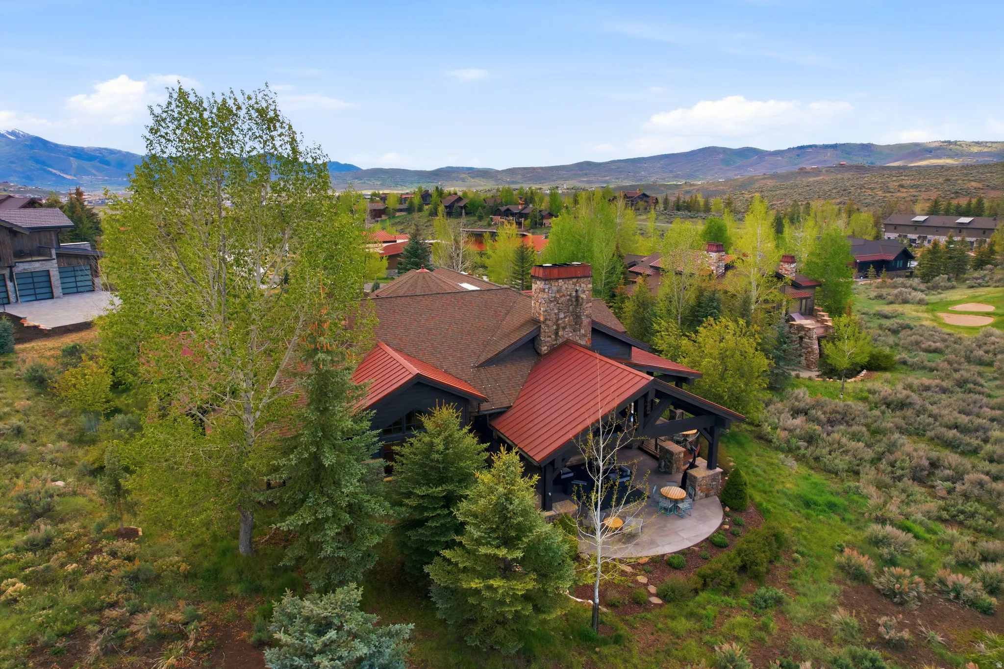 Rustic modern residence featuring a covered patio with a stone base, a stone chimney, and a reddish-brown metal roof accenting a brown shingle roof