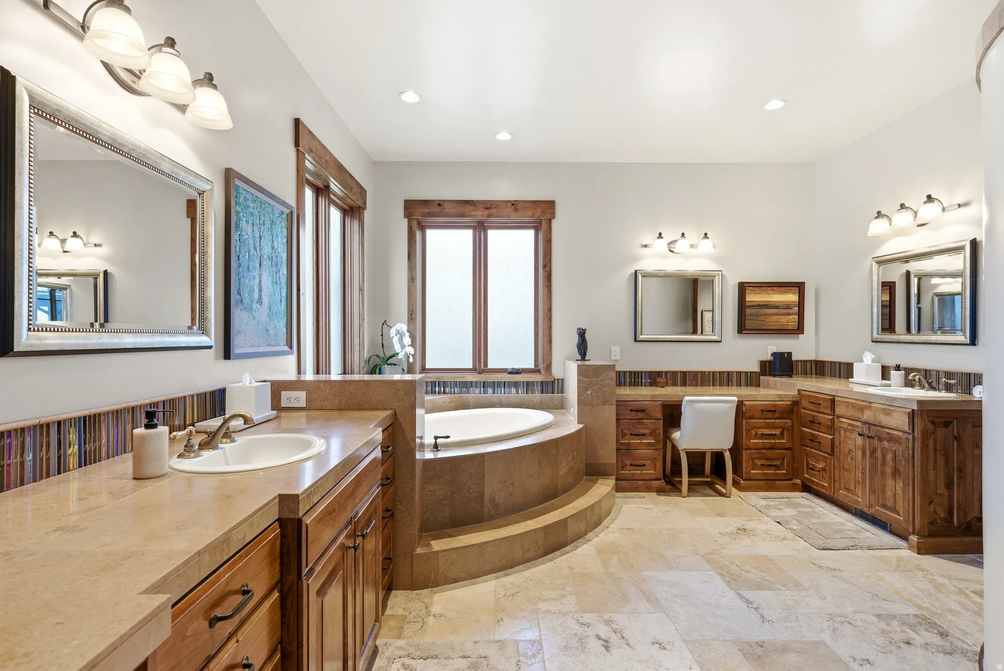 Spacious bath featuring a large built-in soaking tub with tiled surround, extensive wood cabinetry, individual vanity areas, varied tile flooring, and multiple windows with wood trim