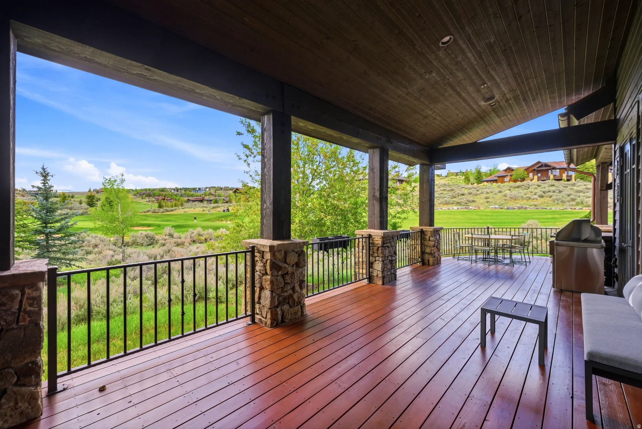 Expansive covered deck featuring rich wood-finish flooring, sturdy stone-wrapped support columns, and a robust black metal railing