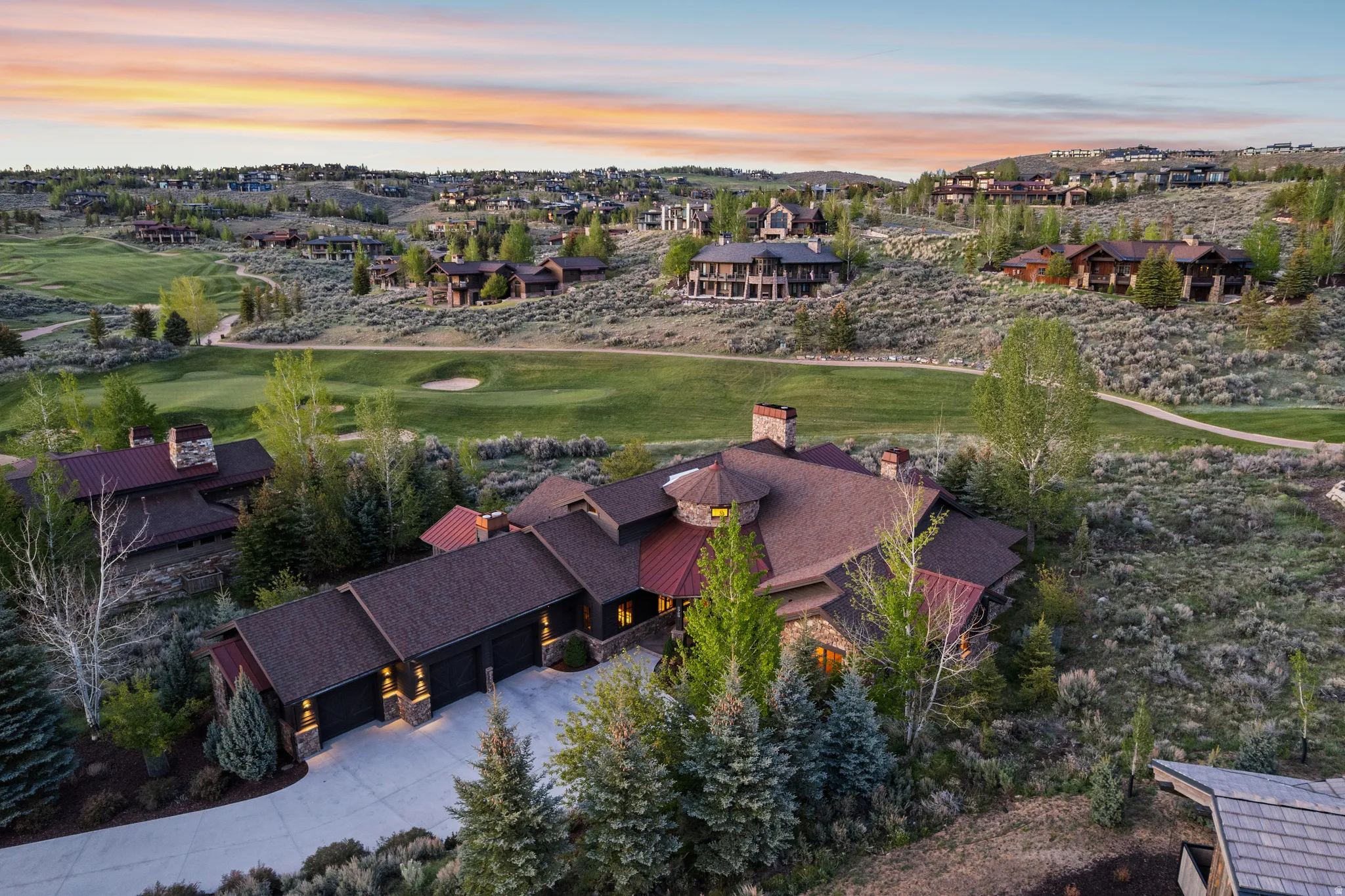 Expansive property featuring a stone and wood facade, a multi-car garage with overhead lighting, a circular turret element, and multiple chimneys, all set within a landscaped hillside with mature trees
