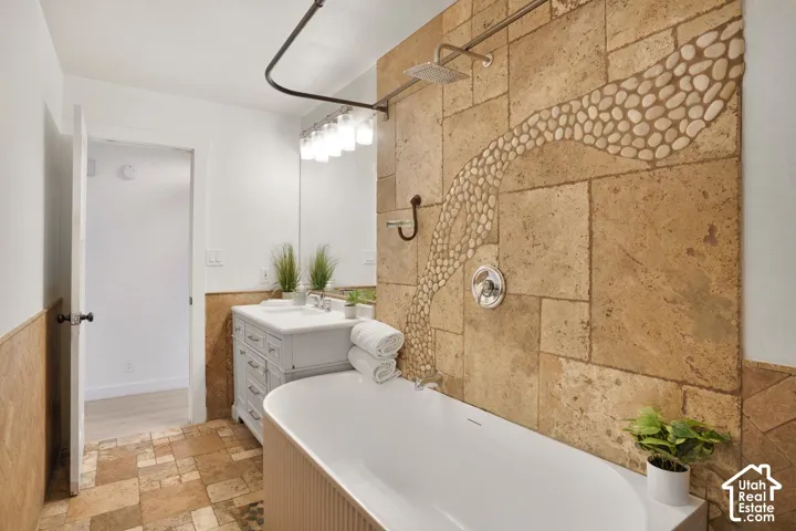 Freestanding soaking tub, stone tile flooring, stone tile shower surround with pebble mosaic, rainfall showerhead, and a vanity with an integrated sink