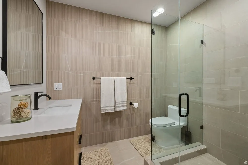Contemporary bathroom featuring a glass-enclosed shower with a matte black handle, a wood-finish vanity with a white countertop and integrated sink, a rectangular framed mirror, and floor-to-ceiling textured wall tile