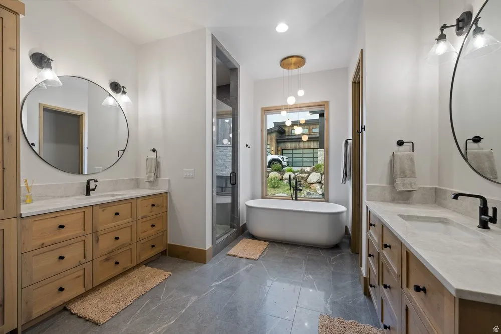 Freestanding soaking tub with a contemporary gold-finish chandelier, dual wood vanities with stone countertops, matte black plumbing fixtures, and a glass-enclosed shower