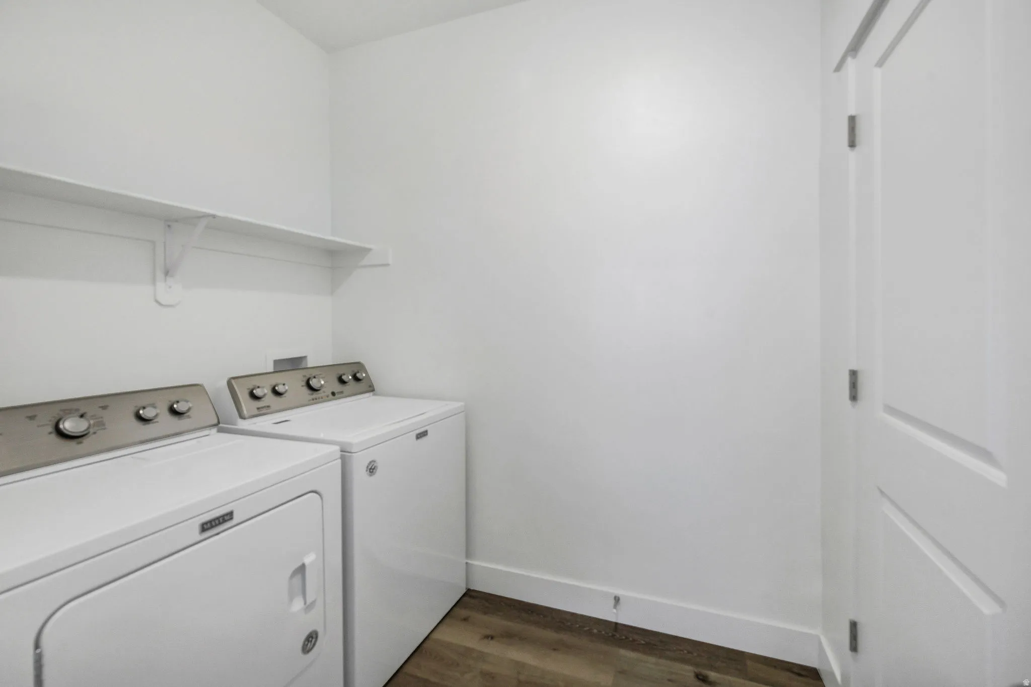 Dedicated laundry area featuring a white floating shelf, neutral white walls, white baseboards, and wood-finish flooring