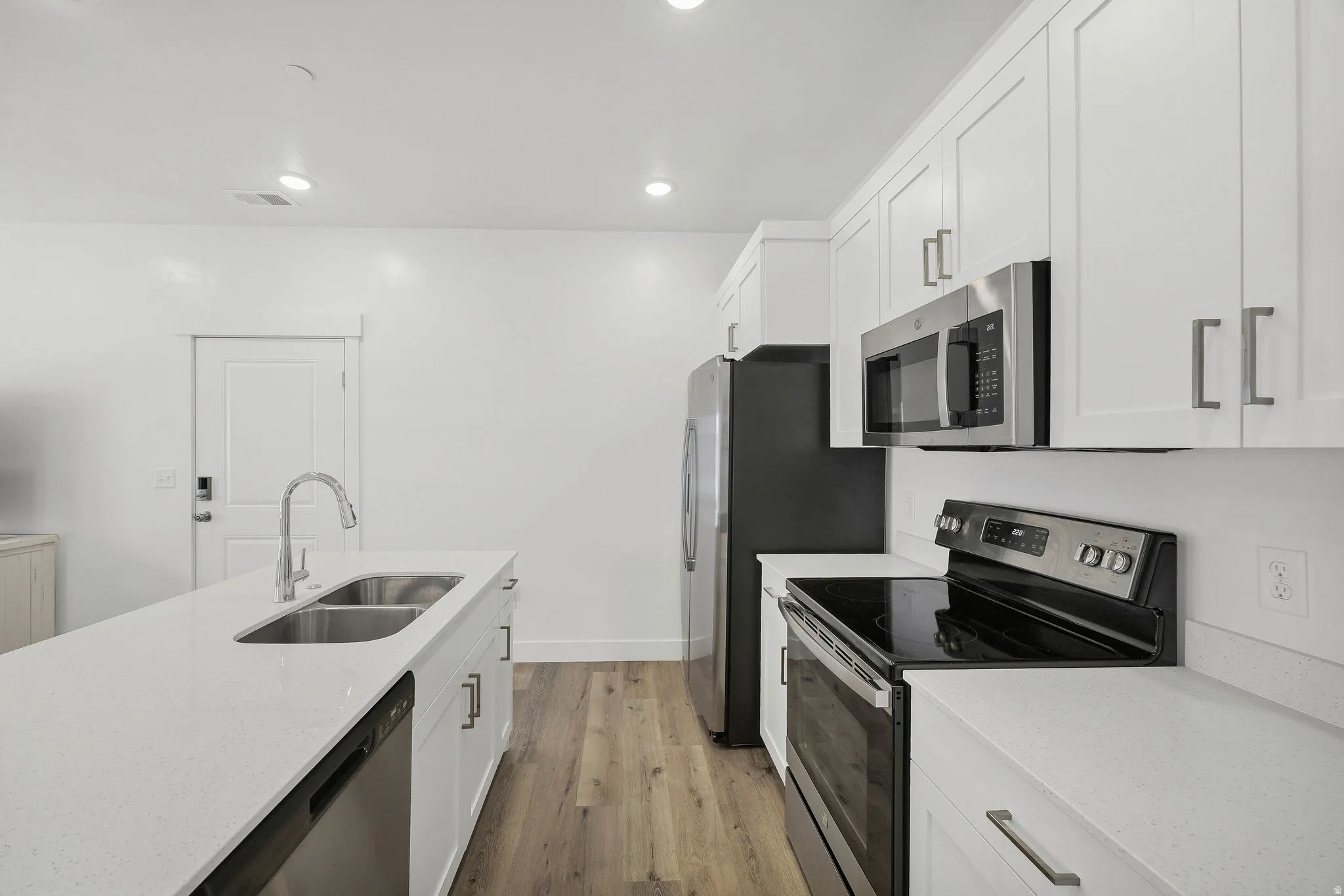 Modern kitchen featuring an island with an undermount double-basin sink, white shaker-style cabinetry, stainless steel appliances, light-toned countertops, and wood-finish flooring