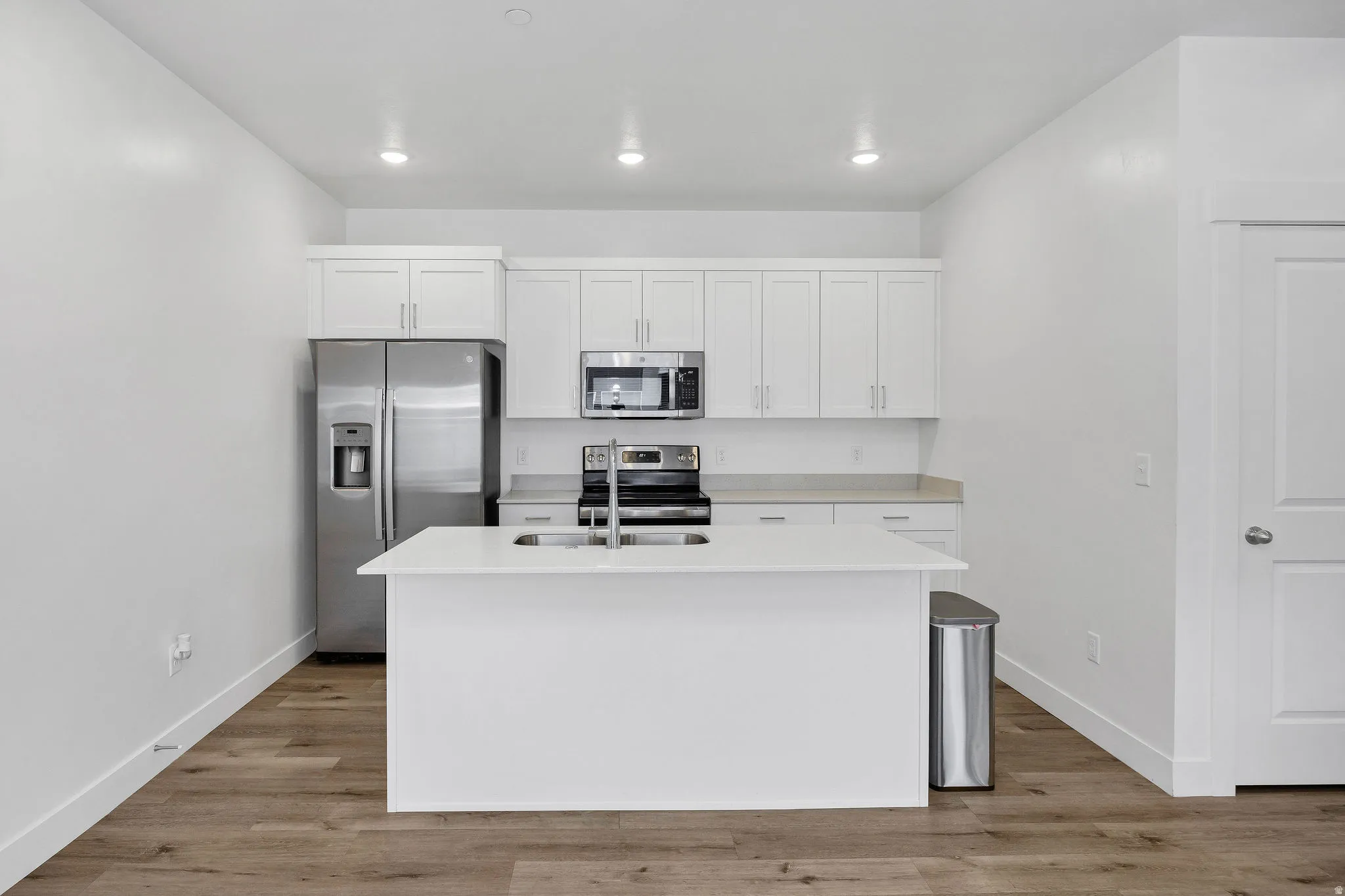 Modern kitchen featuring white shaker cabinetry, stainless steel appliances, a center island with an undermount sink, recessed lighting, and wood-finish flooring