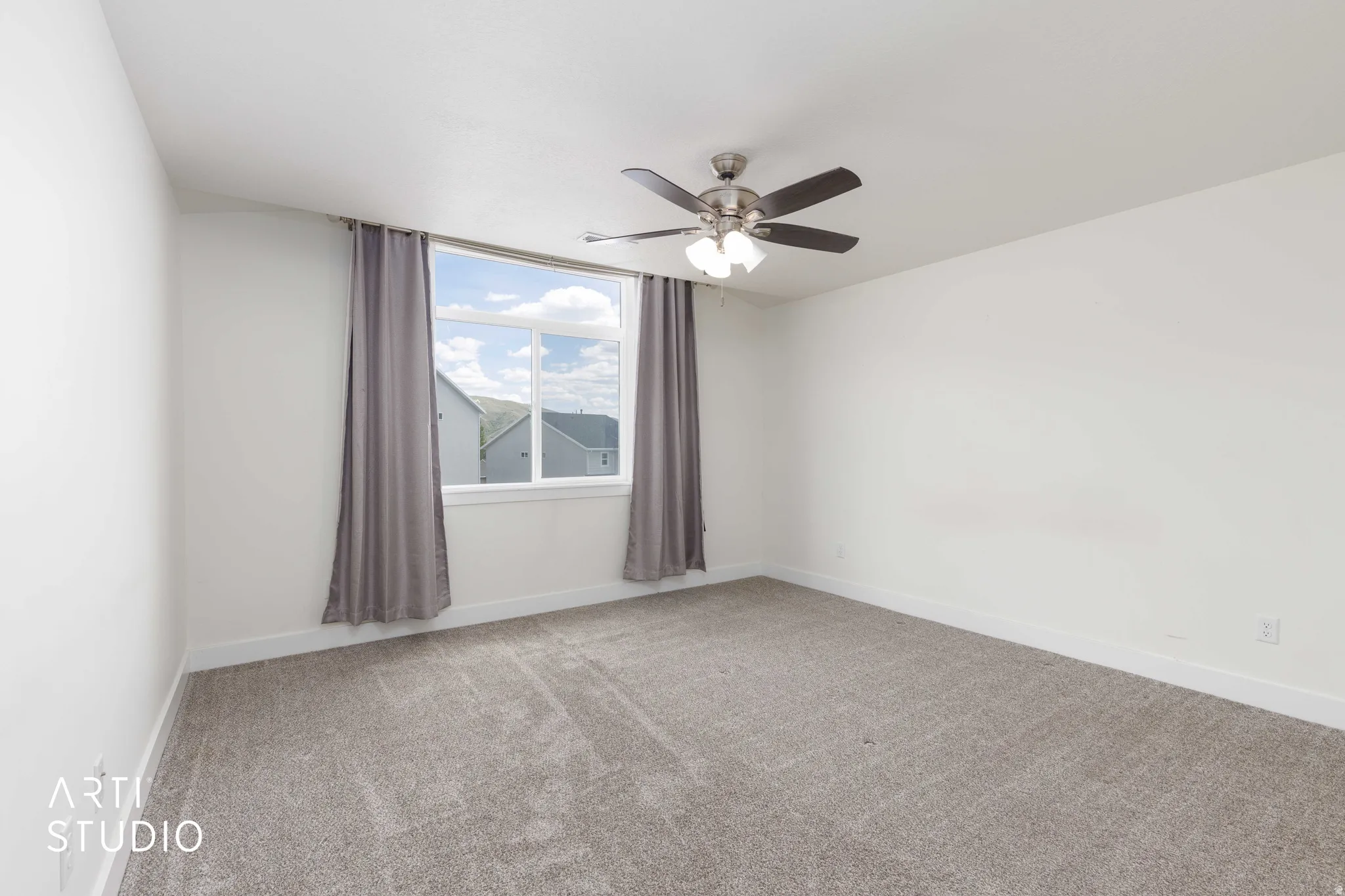 Spacious interior room featuring white walls, grey carpeting, a ceiling fan with integrated lighting, and a large window with grey drapes