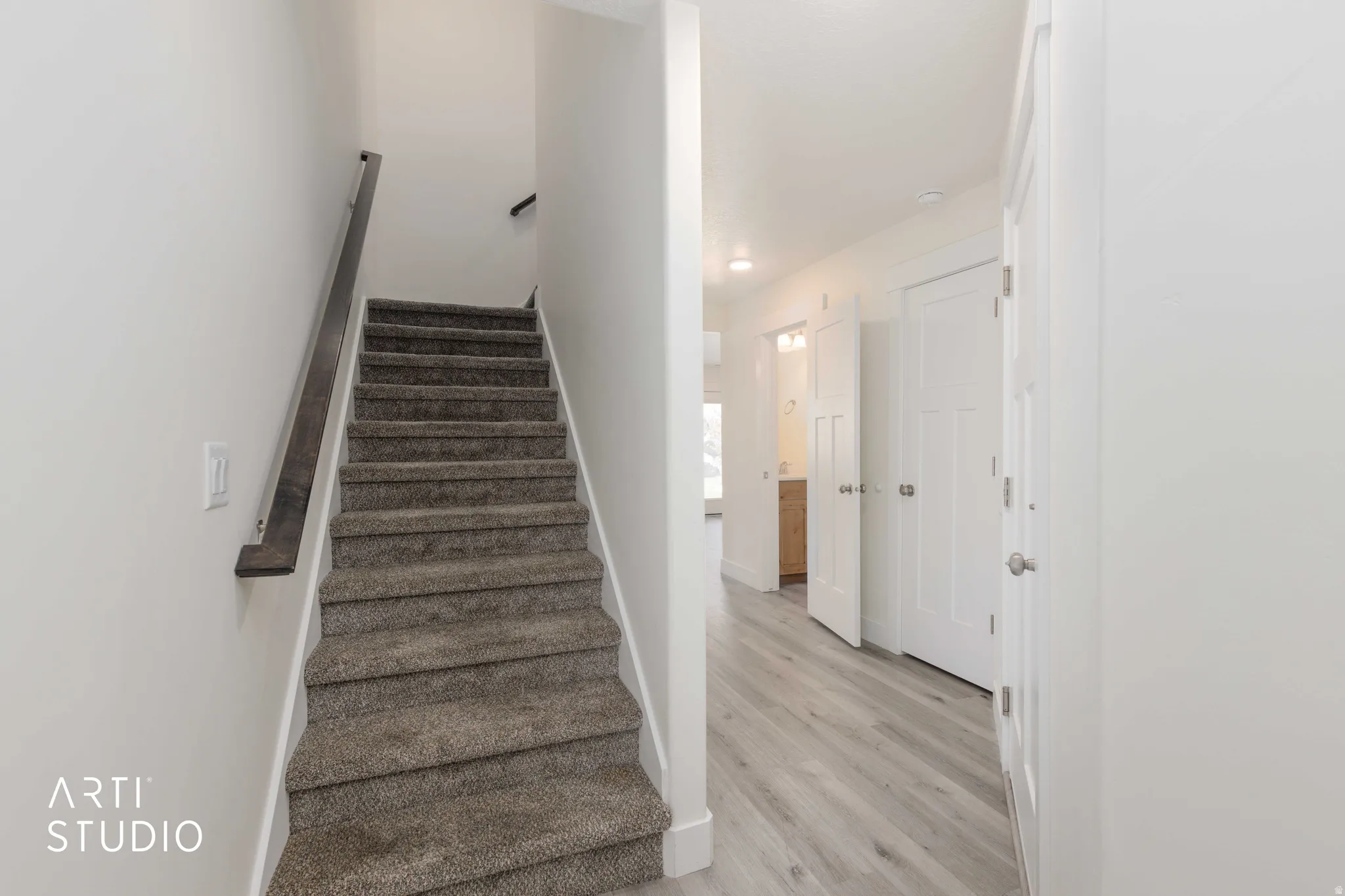 Carpeted staircase with dark wood-finish handrail, light wood-finish flooring, white walls, and white interior doors
