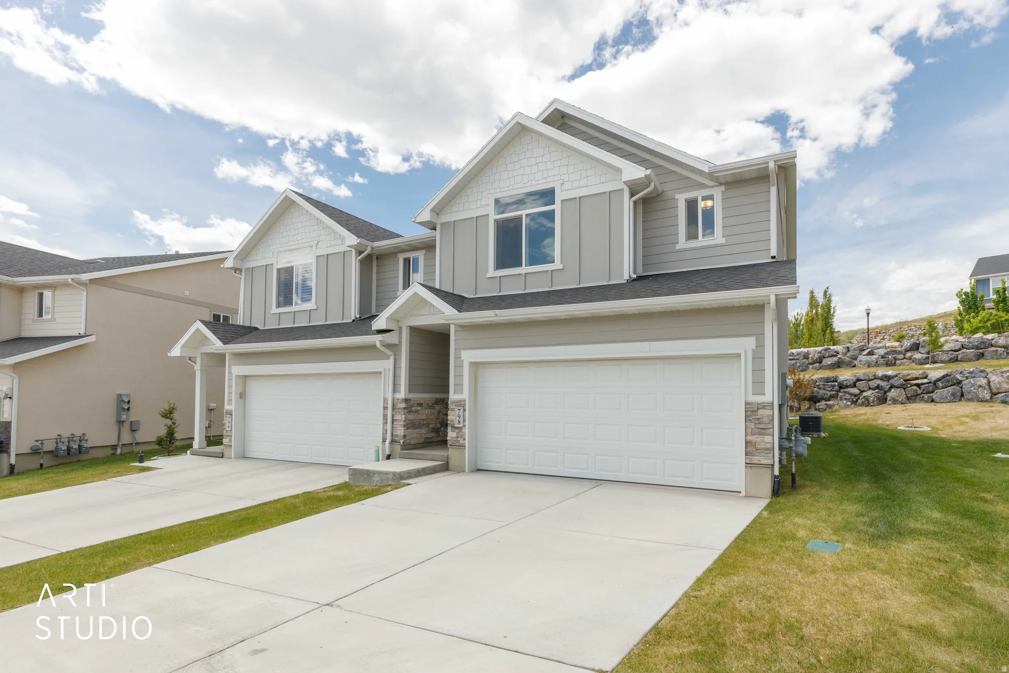 Contemporary townhome exterior featuring light gray siding, white trim accents, prominent gable roofs, attached garages with white panel doors, and stone veneer detailing at the base