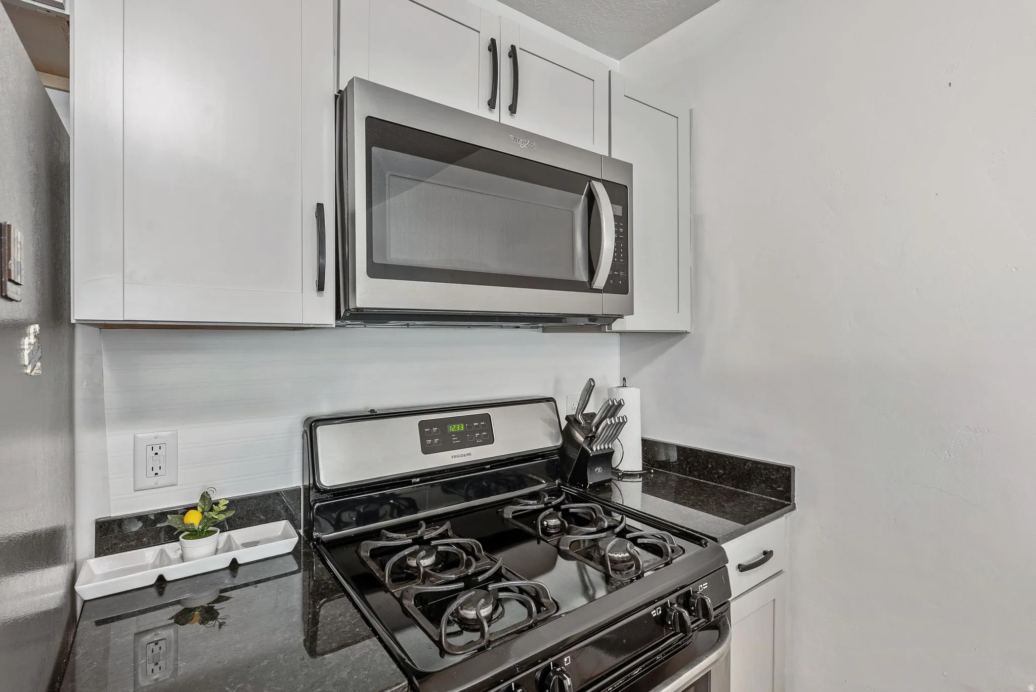 Kitchen featuring a stainless steel microwave, gas range, dark countertops, white cabinetry with dark hardware, and a tile backsplash