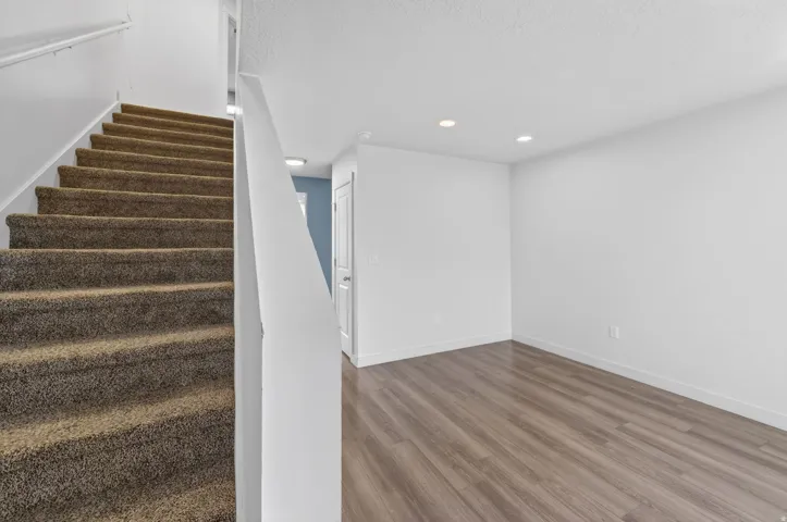 Carpeted staircase featuring a white handrail, contrasting with a room showcasing wood-finish flooring, white baseboards, and recessed lighting