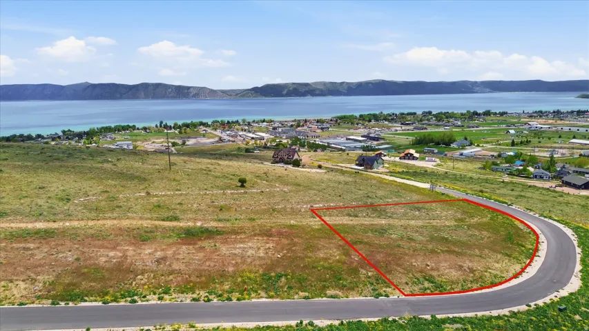 Expansive undeveloped land parcel featuring a paved curved roadway, surrounding natural vegetation, and a prominent lake backdrop