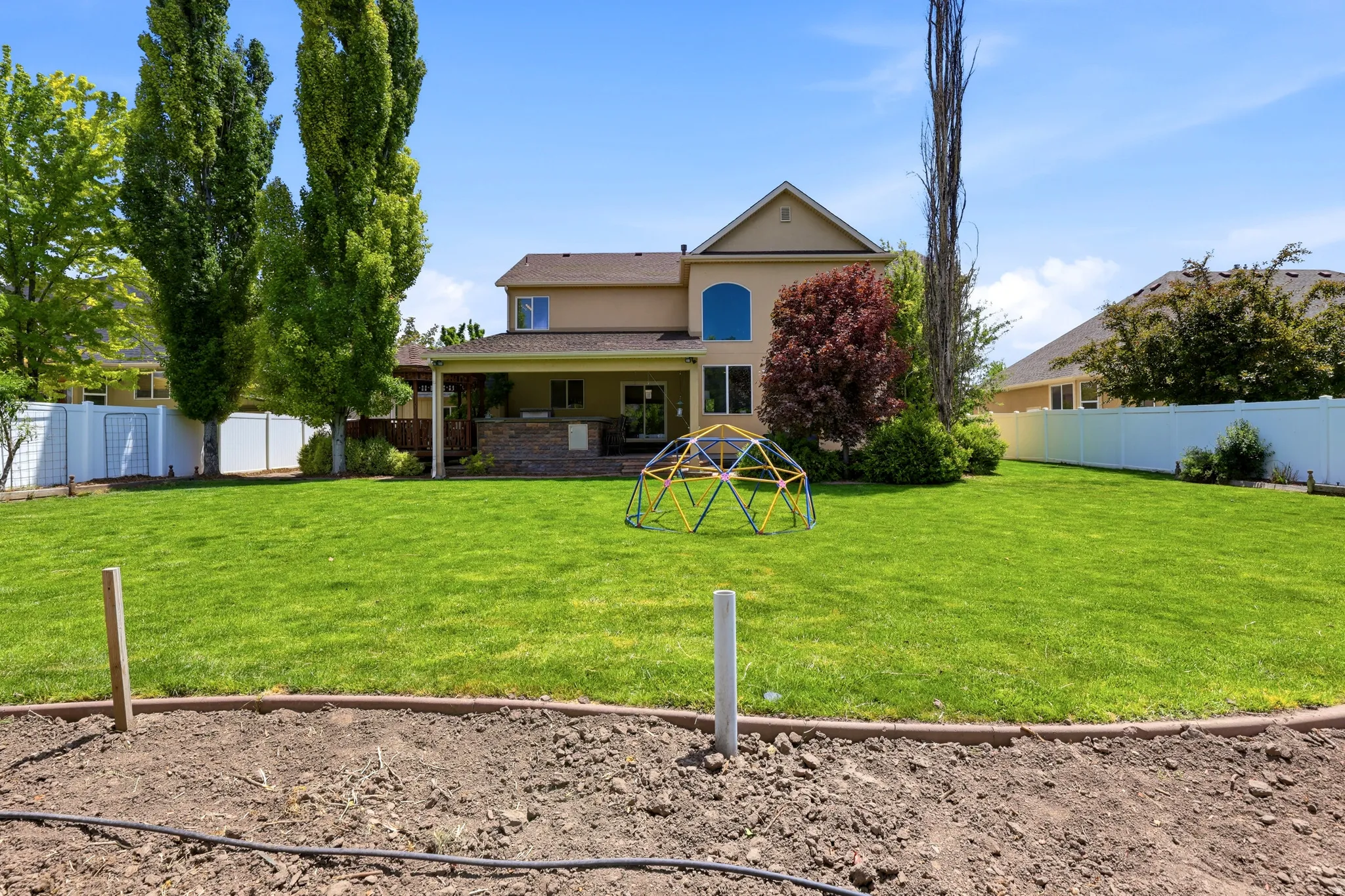 Expansive backyard featuring a manicured lawn, mature trees, and a beige exterior with a covered patio