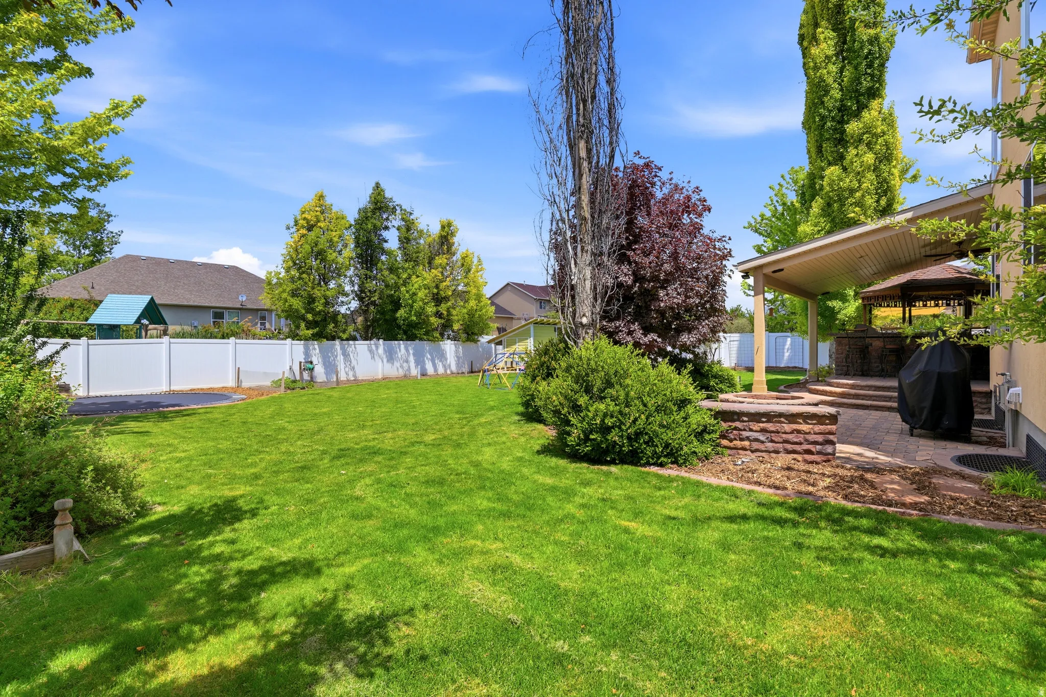 Expansive lawn area featuring a white privacy fence, mature trees, a covered patio, and a built-in brick fire pit