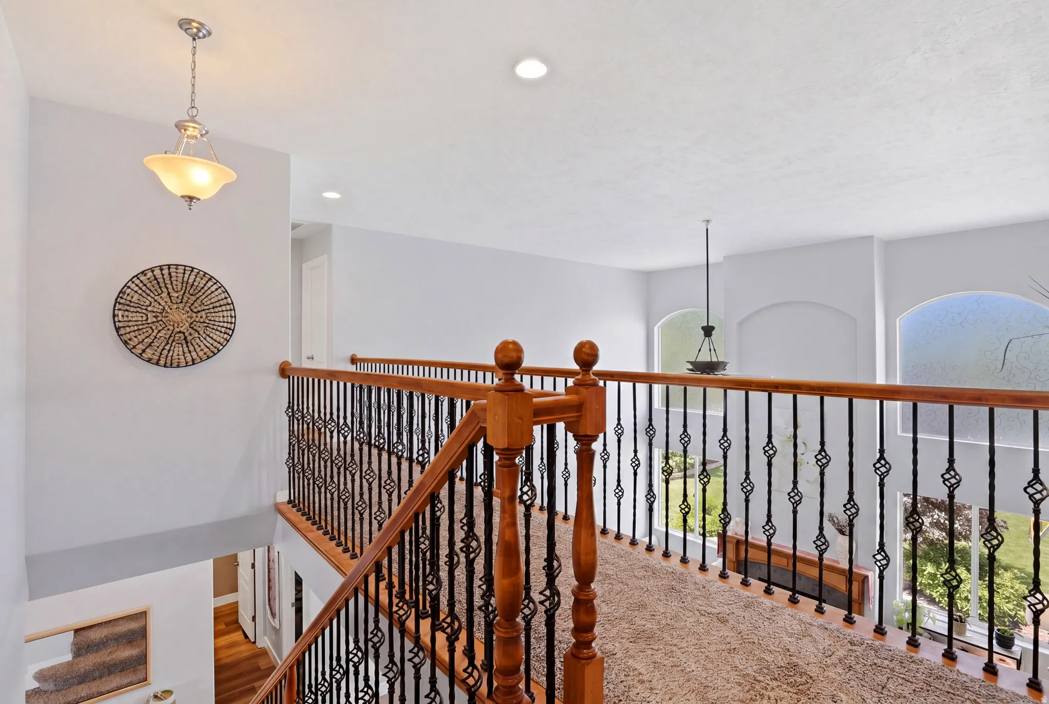 Upper level landing featuring light wood railings with decorative metal balusters, recessed lighting, and a neutral carpeted floor