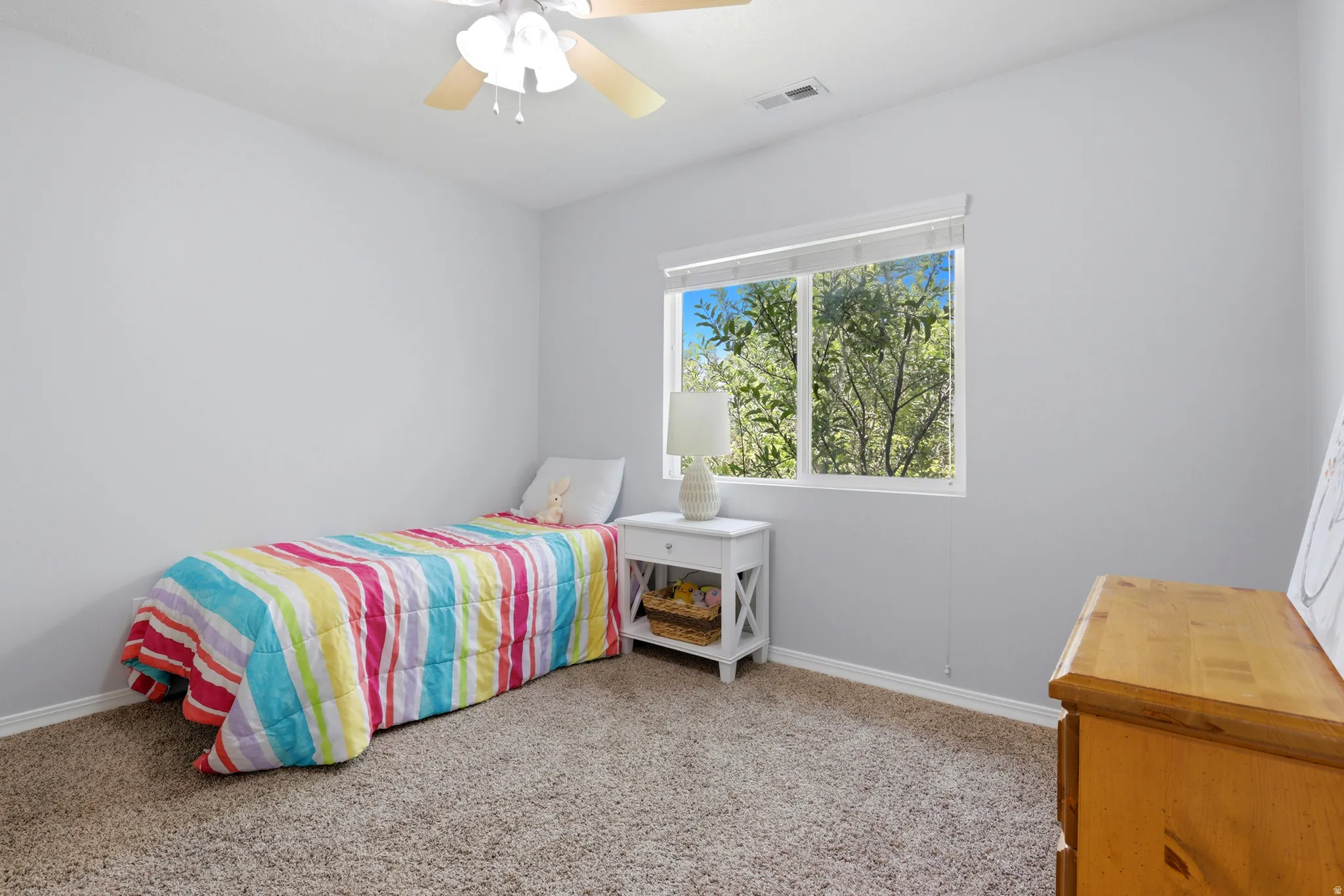 Bright room featuring a window with exterior tree views, neutral wall color, textured carpet flooring, and a ceiling fan with light fixture