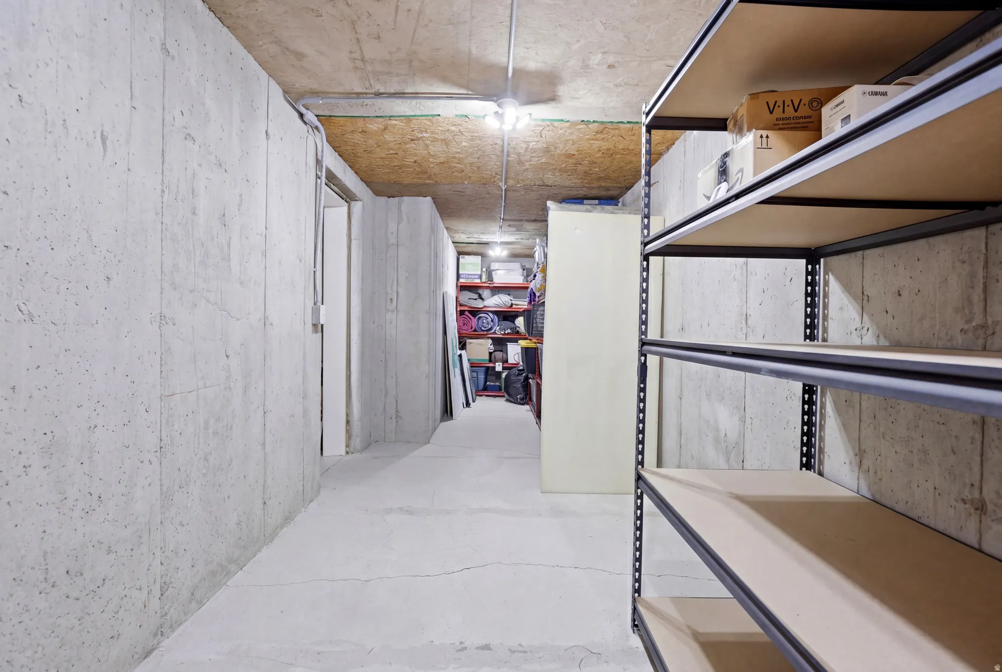 Unfinished basement area featuring concrete walls and flooring, exposed ceiling joists, and built-in metal shelving units