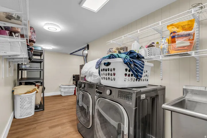 Dedicated laundry space featuring wood-finish flooring, two large capacity laundry appliances, a stainless steel utility sink, and extensive wall-mounted shelving