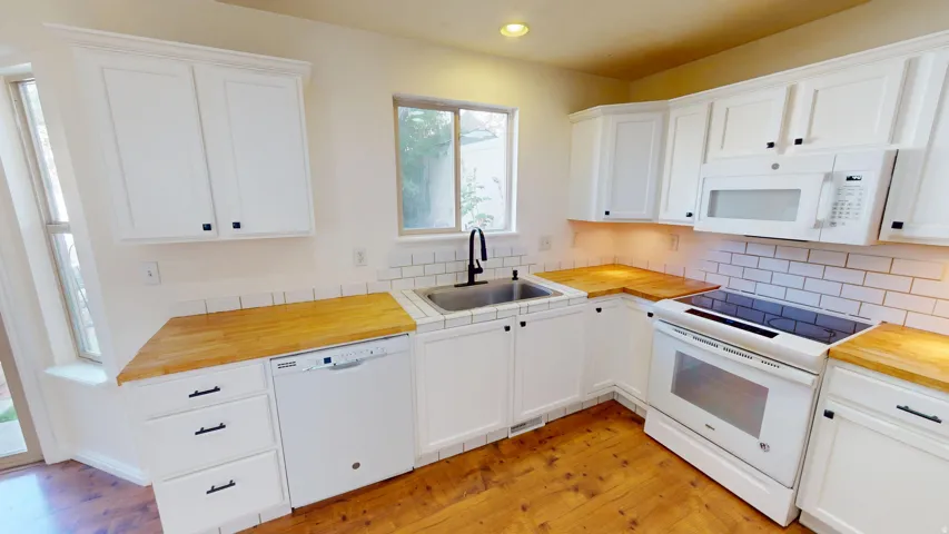 White cabinetry with matte black hardware, wood-finish countertops, white subway tile backsplash, stainless steel sink, and wood-finish flooring