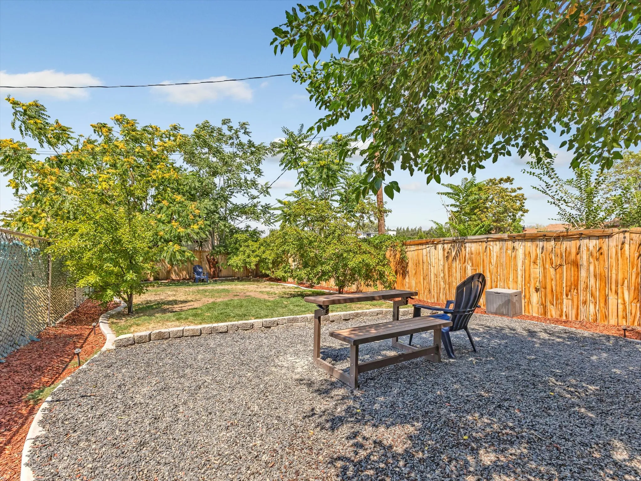 Spacious backyard featuring a dedicated gravel patio area with a wooden picnic bench, a grassy lawn, mature trees providing natural shade, a wood privacy fence, and a chain-link fence