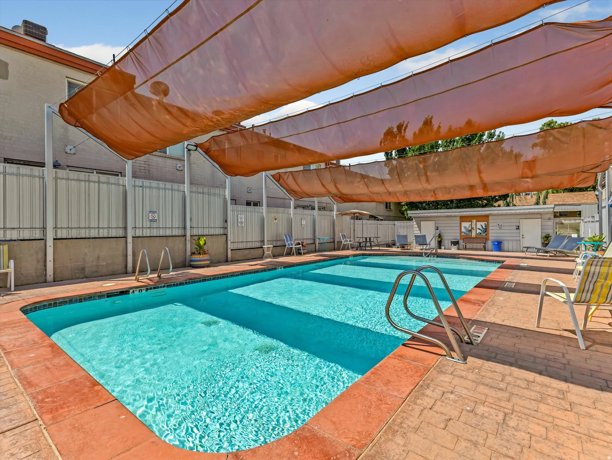 Rectangular swimming pool with brick coping and stamped concrete decking, featuring a stainless steel pool ladder, surrounding privacy fencing, and overhead shade canopies