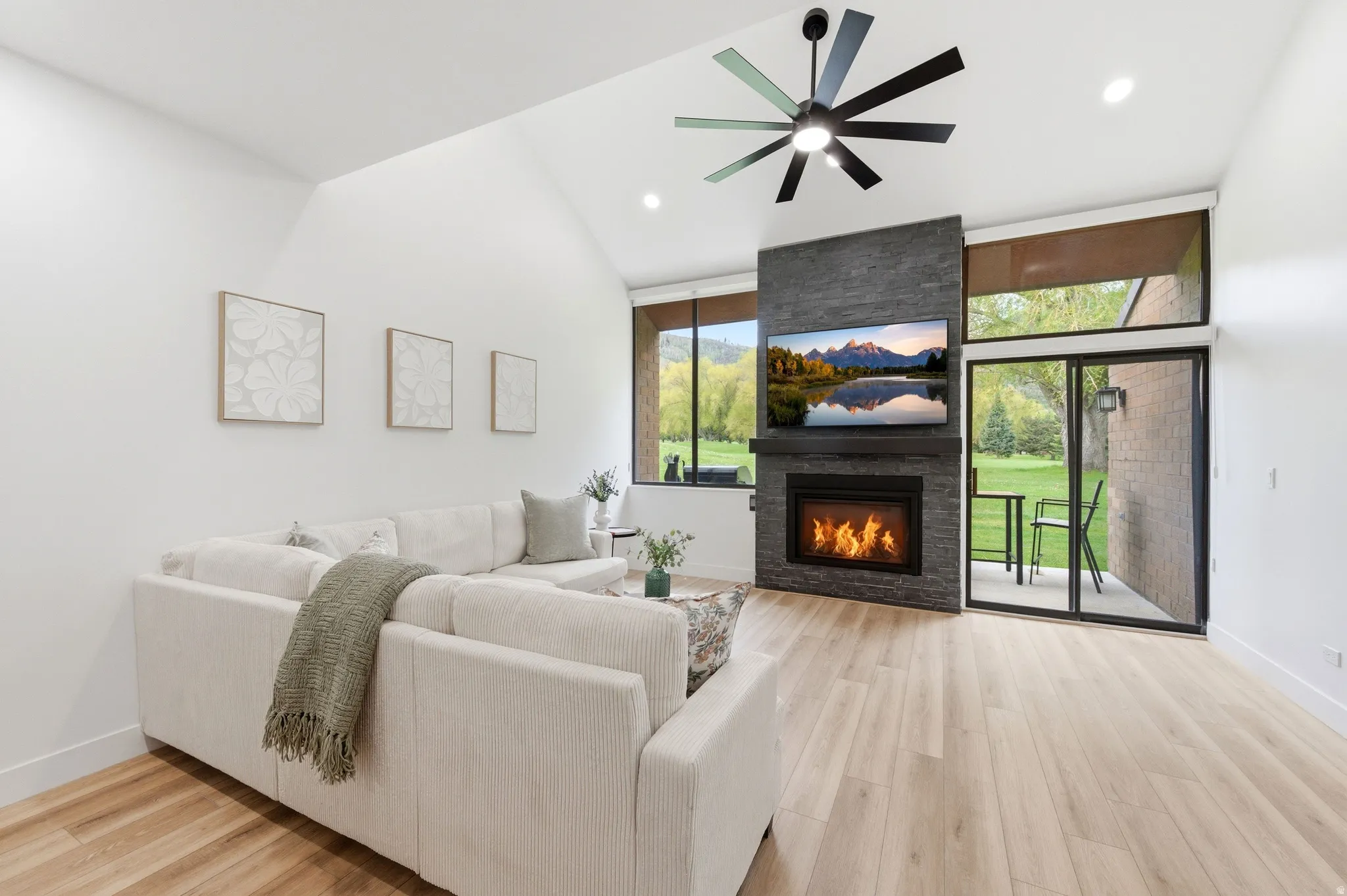 Vaulted ceiling with a contemporary ceiling fan, stone-clad fireplace with a built-in mantel, wood-finish flooring, full-height windows, and a glass sliding door leading to an outdoor patio area