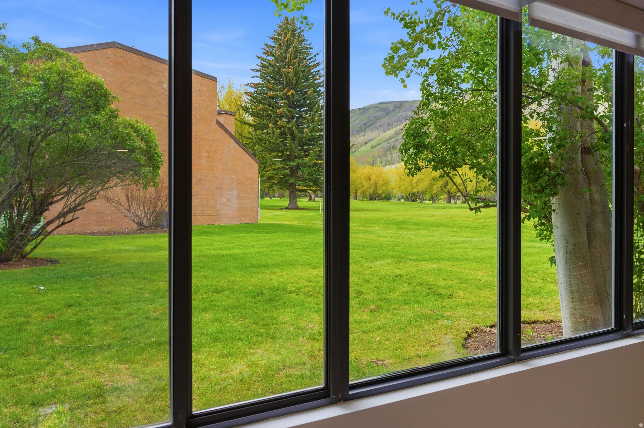 Floor-to-ceiling windows with dark frames, showcasing a verdant lawn, mature trees, distant mountains, and a brick-clad building