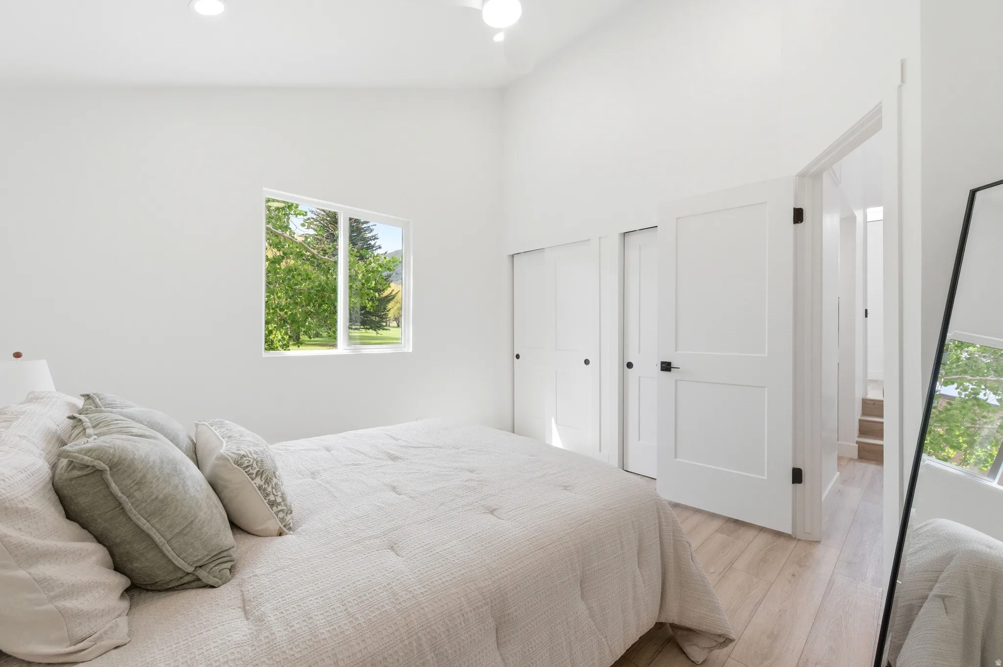 Light-filled bedroom featuring a window with green outdoor scenery, vaulted ceilings, recessed lighting, wood-finish flooring, and two panel doors with matte black hardware