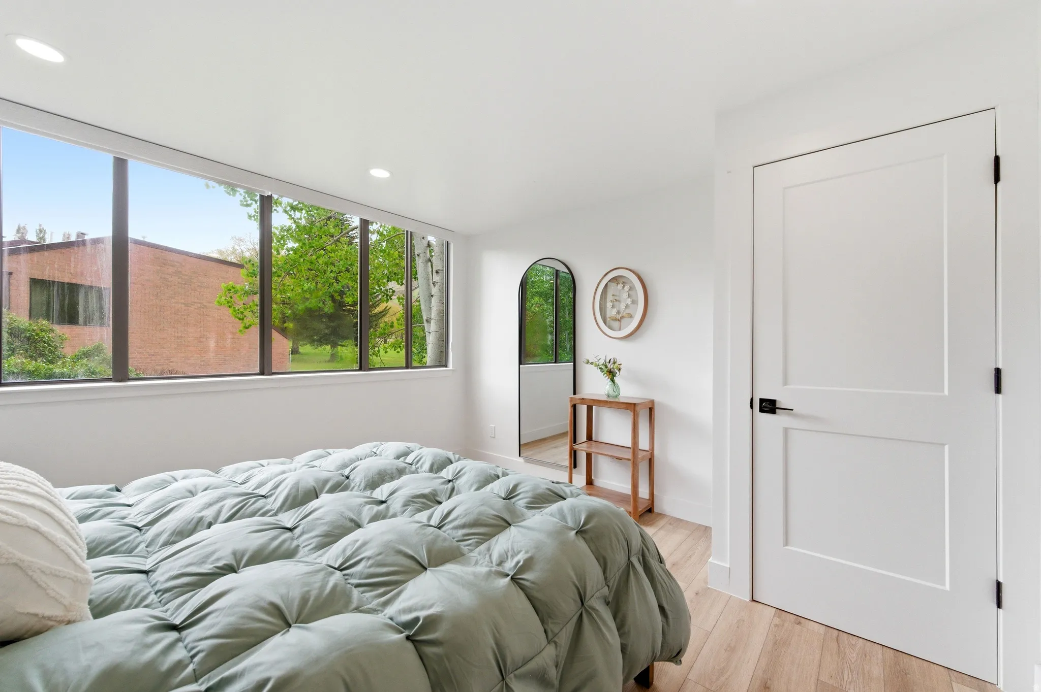Expansive window wall with dark frames, recessed ceiling lighting, light wood-finish flooring, and a white two-panel interior door with matte black hardware