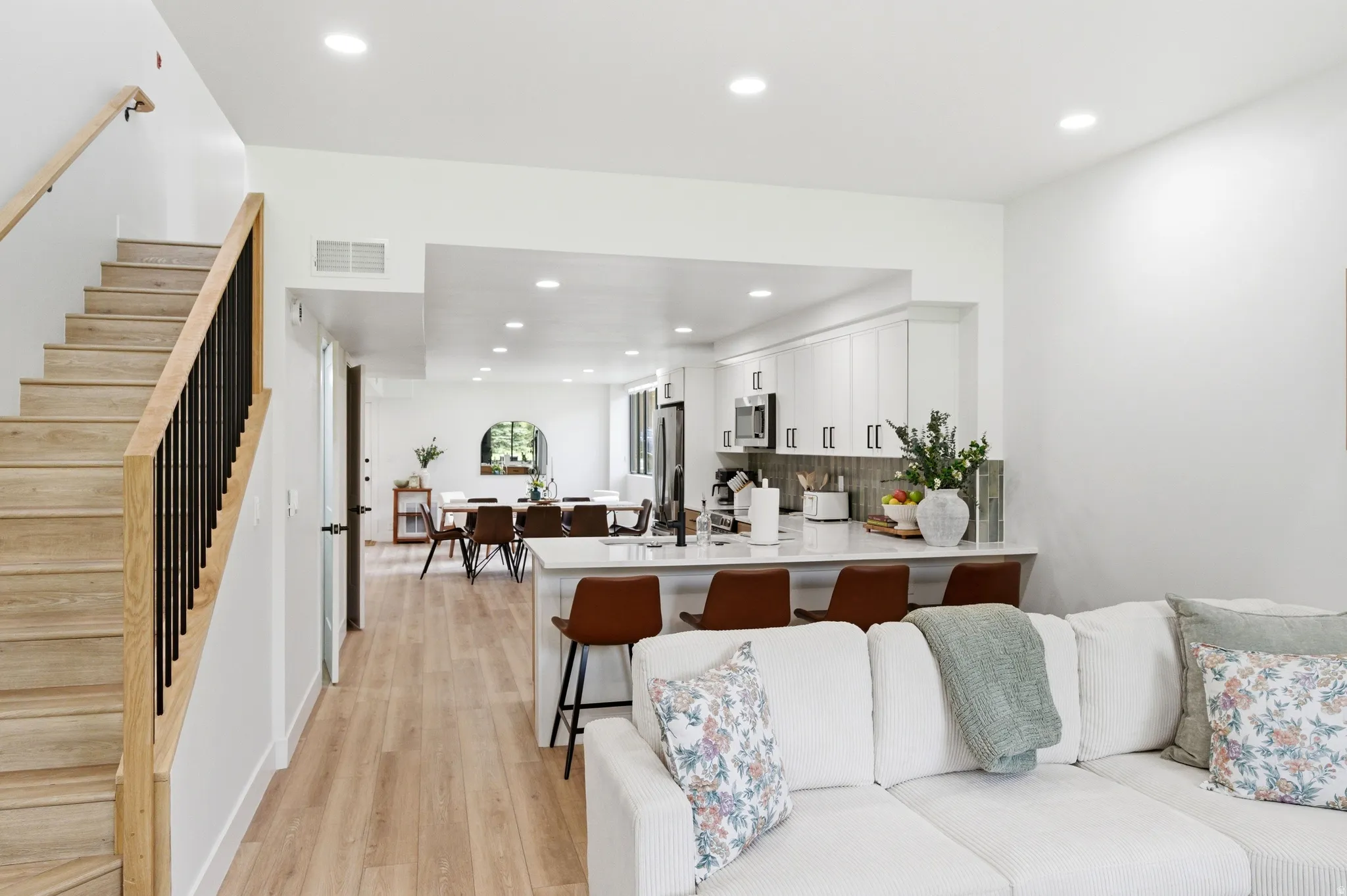 Open-concept living space featuring wood-finish flooring, a contemporary staircase with natural wood handrail and black metal balusters, recessed lighting, white cabinetry, and a kitchen island with seating