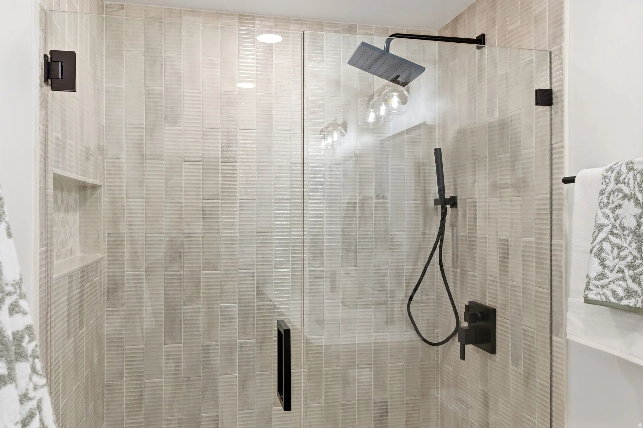 Glass-enclosed shower featuring vertically stacked tile, a built-in shower niche, and matte black fixtures including a rainfall shower head and handheld sprayer