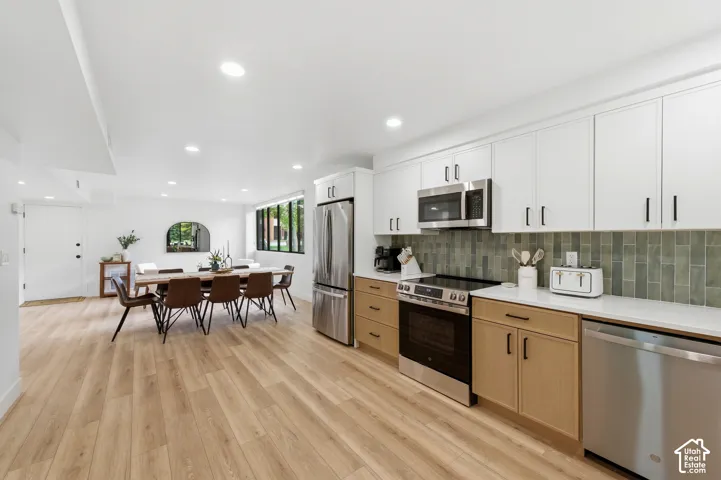 Modern open-concept kitchen and dining area featuring white cabinetry with brushed metal hardware, wood-finish lower cabinets, stainless steel appliances including a built-in microwave, quartz countertops, and a green subway tile backsplash