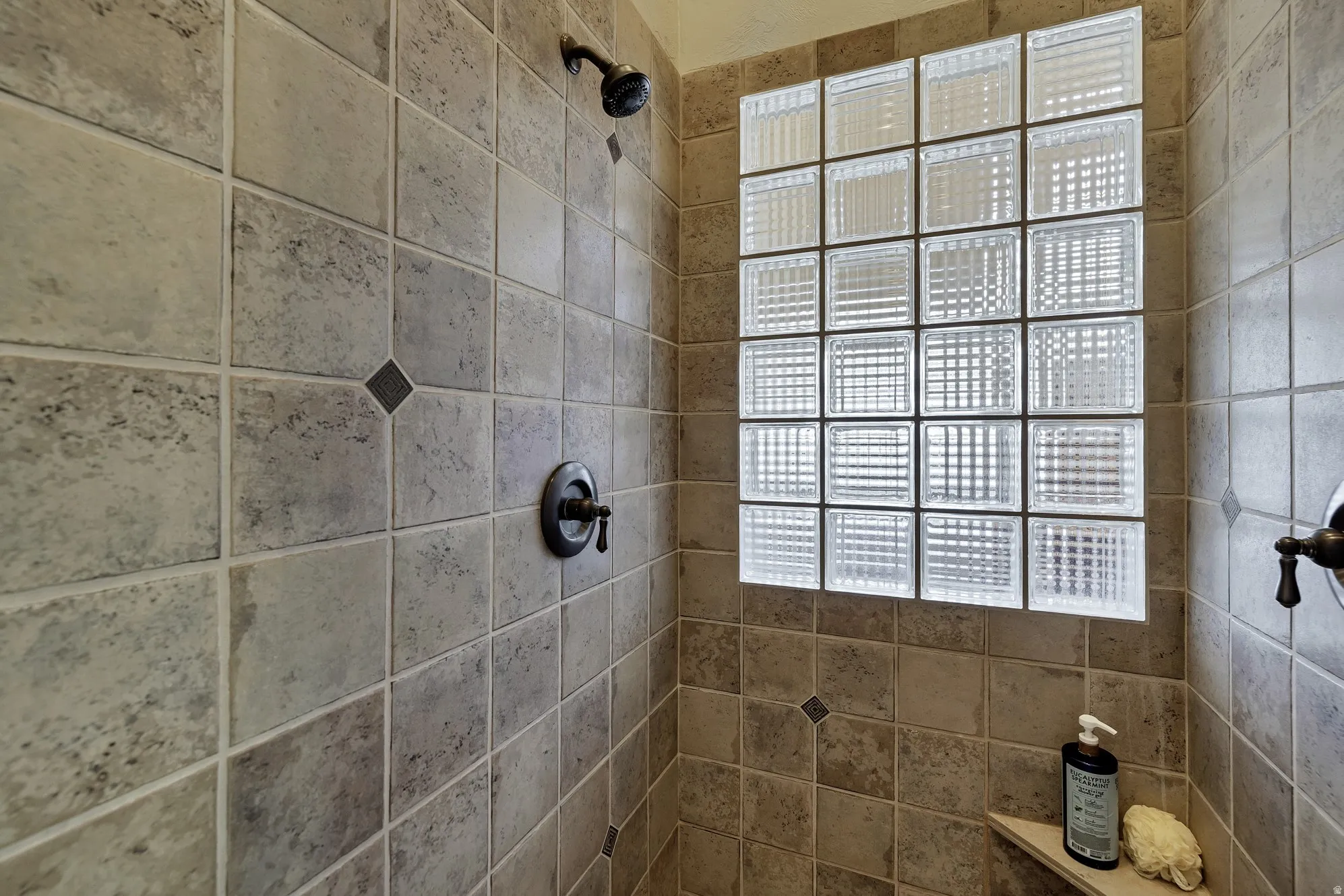 Shower enclosure featuring textured wall tiling with accent tiles, a glass block window, oil-rubbed bronze fixtures, and a corner shelf