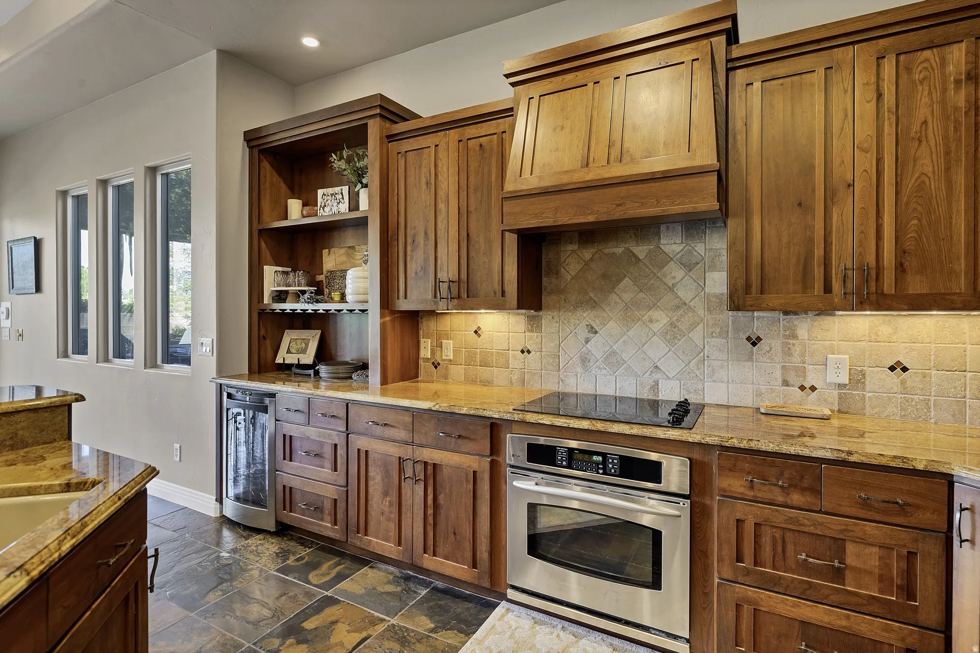 Gourmet kitchen featuring rich wood cabinetry, granite countertops, stainless steel wall oven, cooktop, and a tile backsplash with decorative accents
