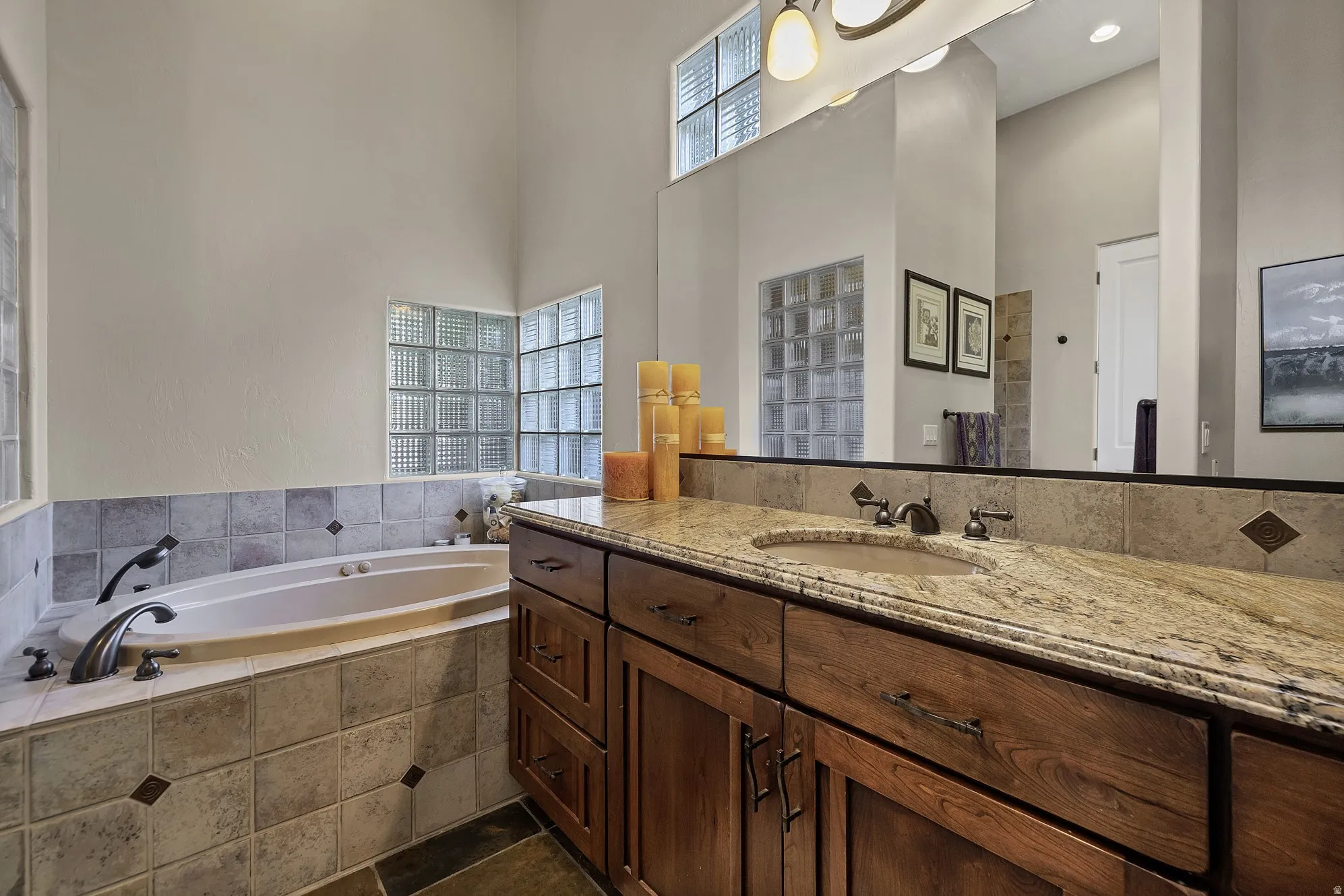 Spacious bath featuring a built-in soaking tub with tiled surround, dual vanity with granite countertop, and glass block windows