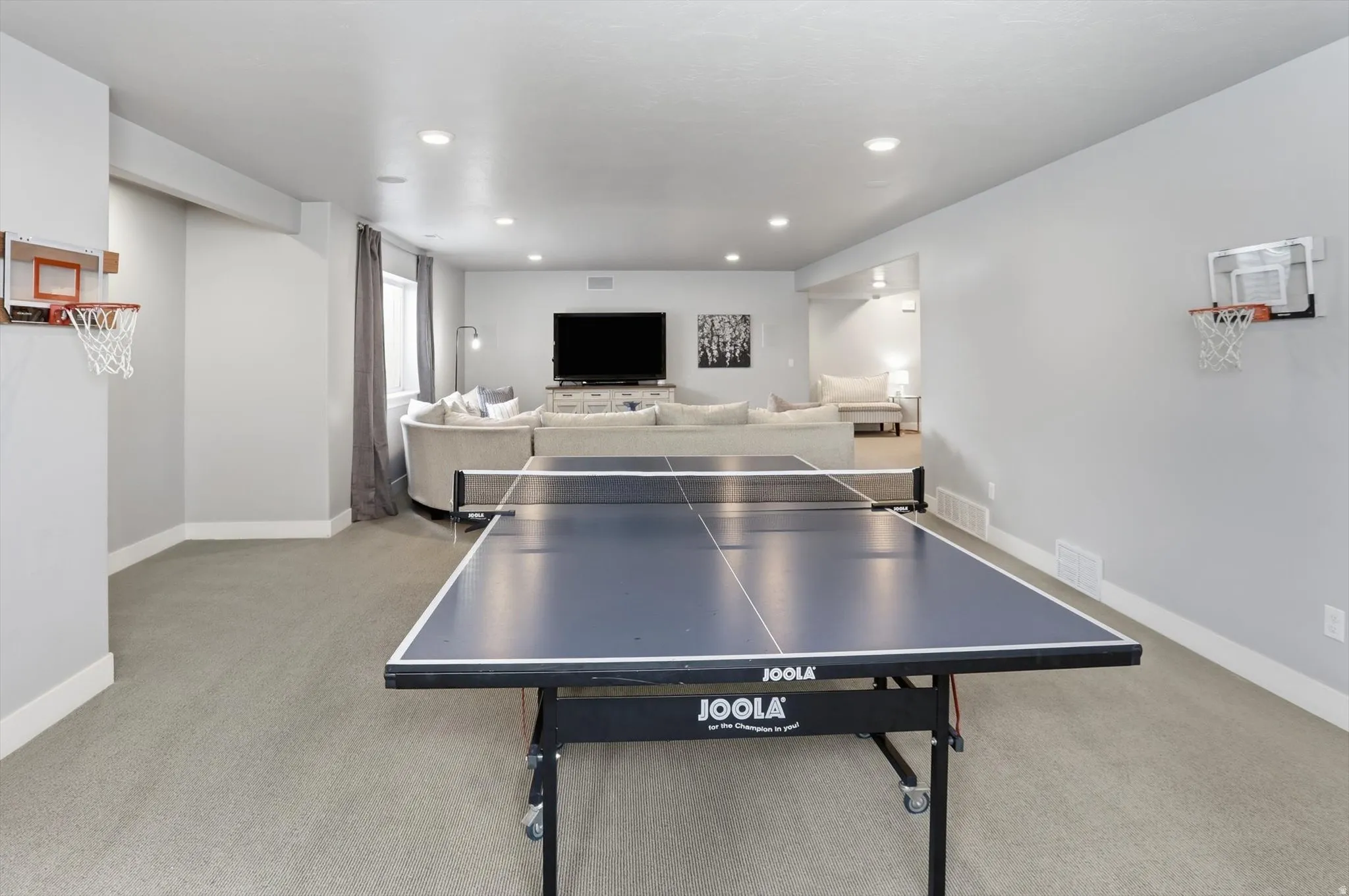Spacious recreation area featuring light gray carpeting, recessed lighting, and a large window with gray drapes