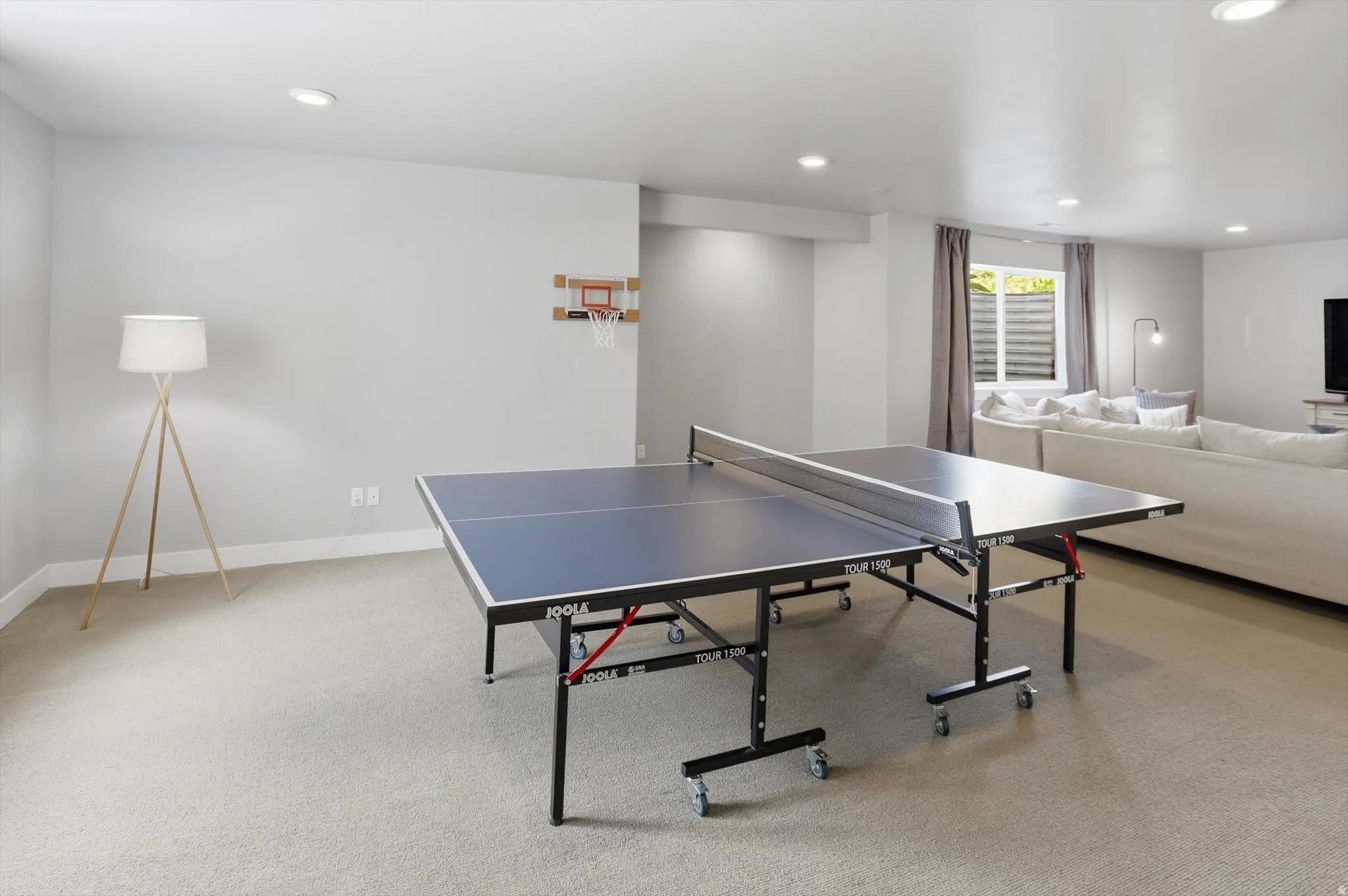 Spacious recreation room featuring light-toned carpeting, neutral wall finishes, recessed lighting, and a large window with privacy blinds