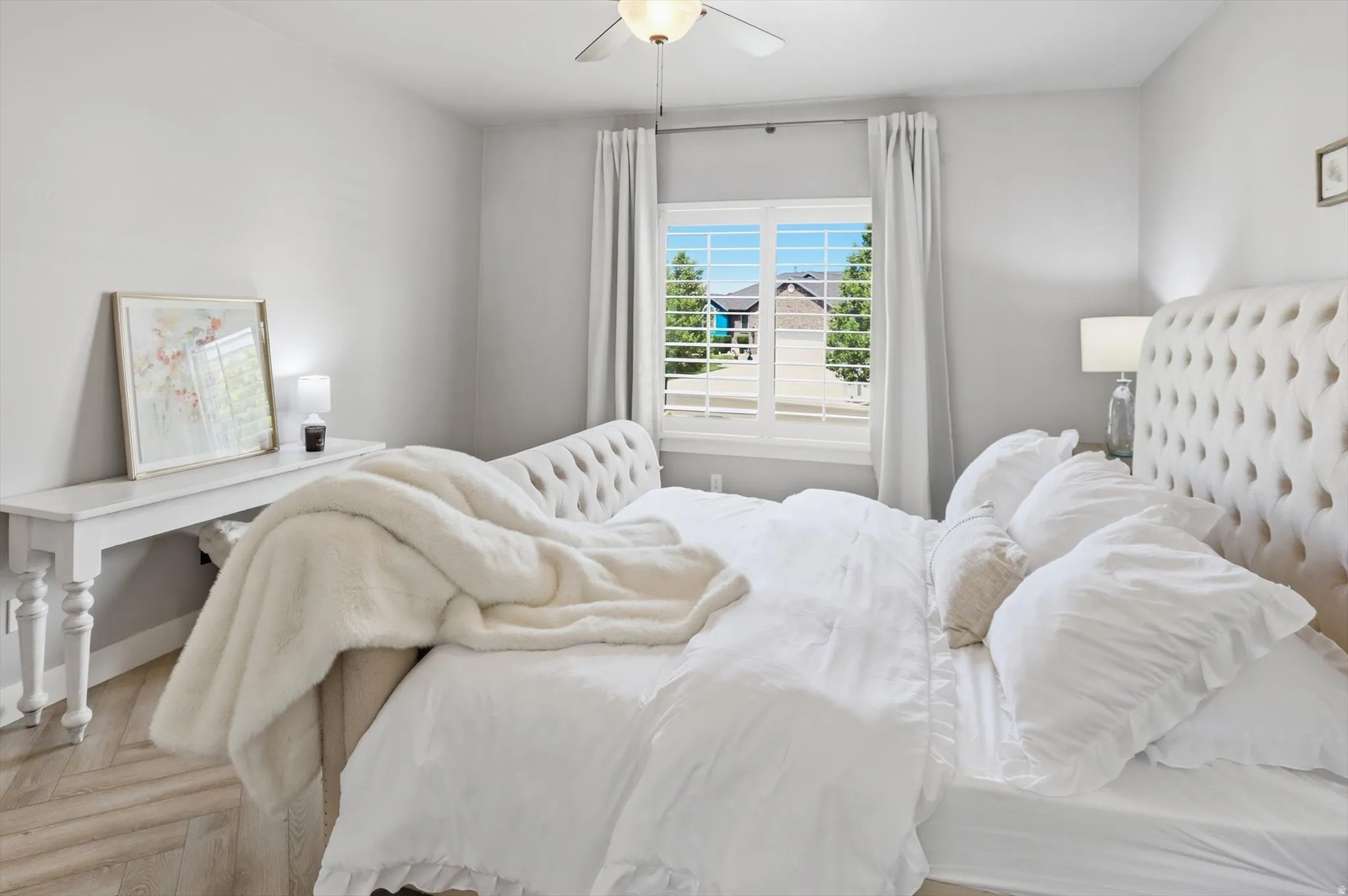 Bedroom featuring light wood-finish flooring, a white window with plantation shutters, ceiling fan with integrated lighting, and neutral wall paint