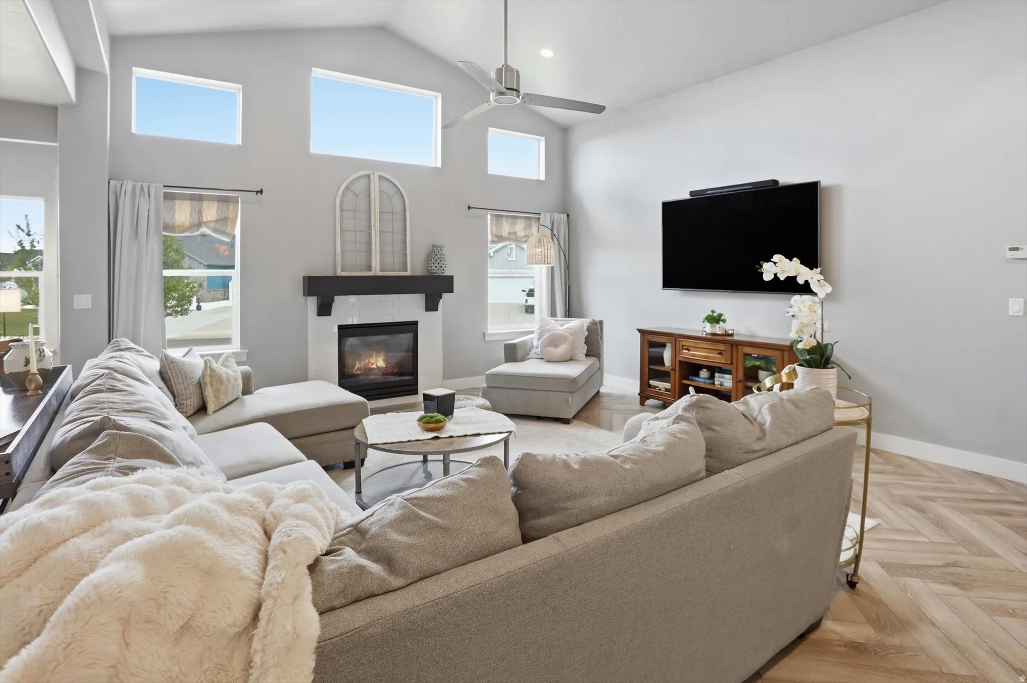 Living area featuring a vaulted ceiling with a fan, a fireplace with a dark wood mantel and white tile surround, multiple windows, and wood-finish flooring with a herringbone pattern