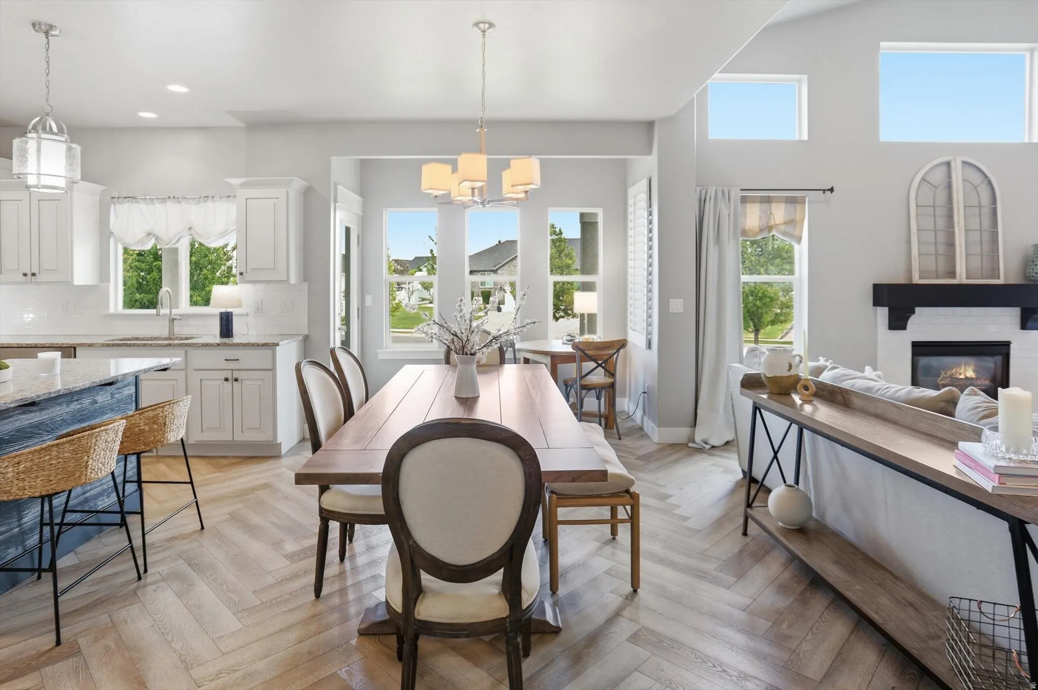 Open-concept living area featuring herringbone-patterned wood-finish flooring, a white-tiled fireplace with a dark wood mantel, and abundant natural light from multiple windows