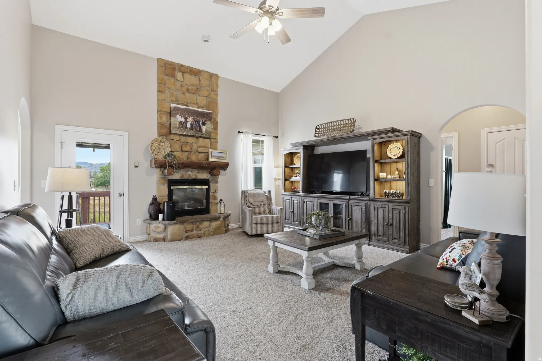 Vaulted ceiling living area featuring a stone fireplace with wood mantel, light-toned carpeting, an arched doorway, and outdoor access through a glass-paneled door