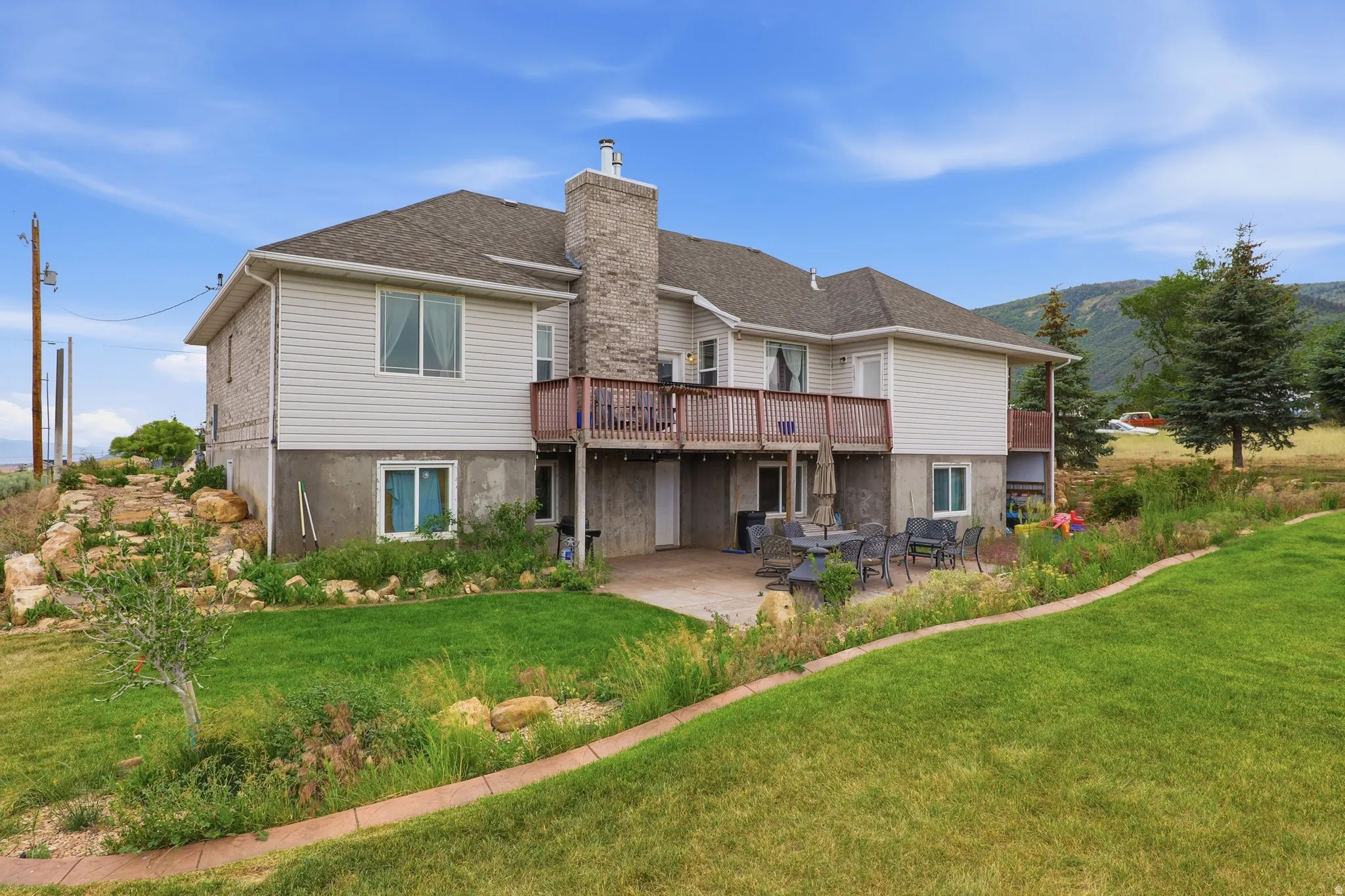 Rear exterior featuring a stone chimney, upper-level wood deck, lower-level concrete patio, and a sloped lawn