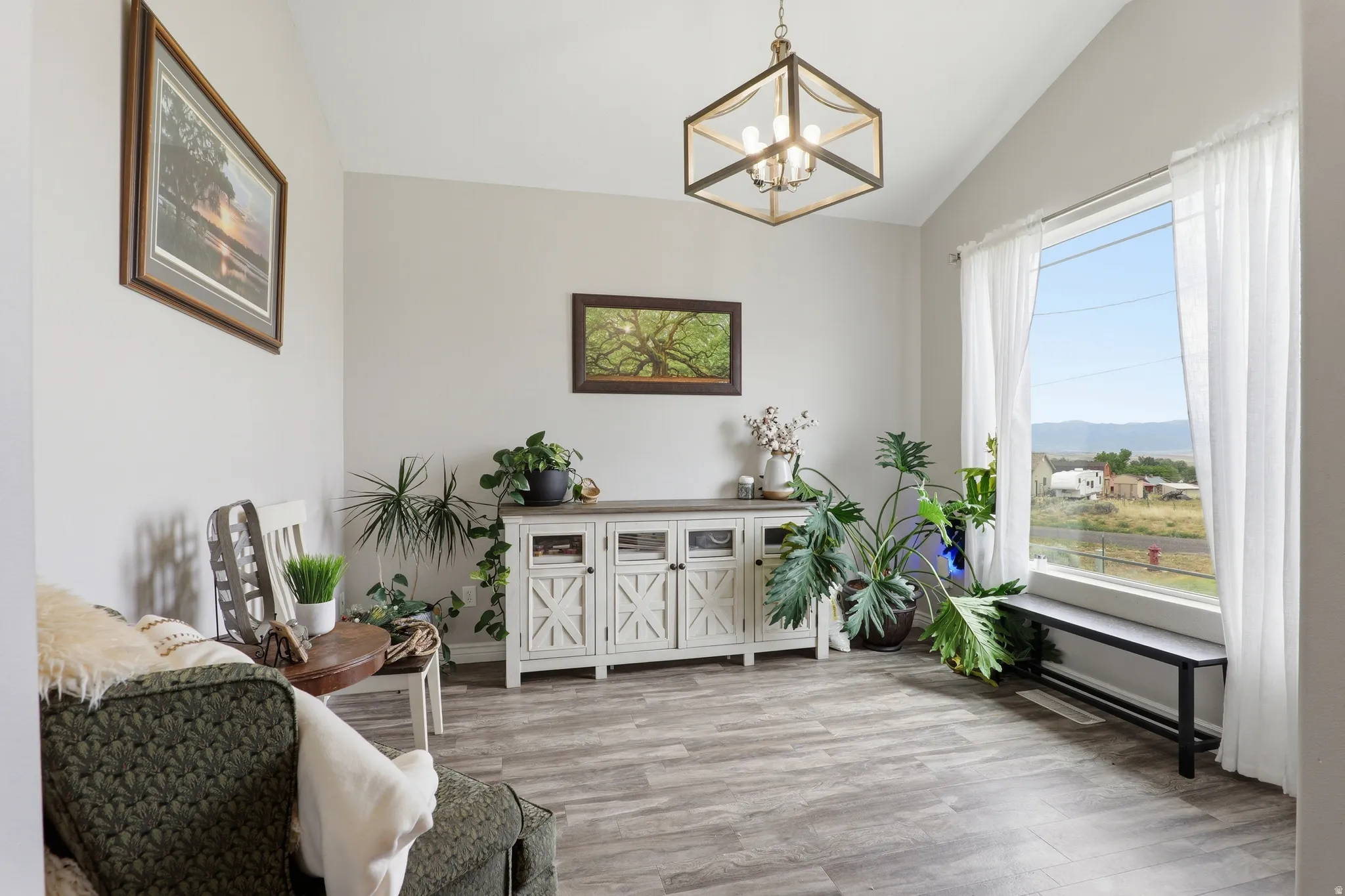 Vaulted ceiling featuring a geometric chandelier, large picture window with white sheer curtains, wood-finish flooring, and a distressed white console table