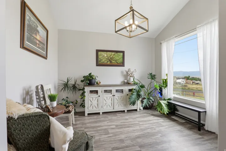 Vaulted ceiling featuring a geometric chandelier, large picture window with white sheer curtains, wood-finish flooring, and a distressed white console table