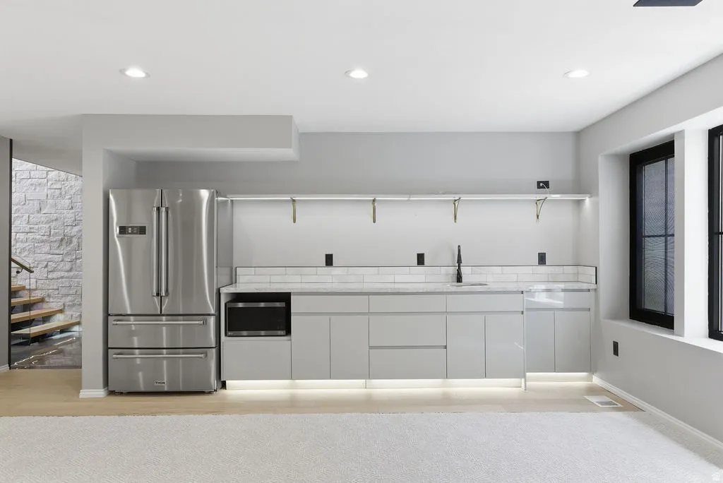 Modern wet bar featuring a stainless steel French-door refrigerator, integrated microwave, undermount sink with a matte black faucet, and light gray cabinetry with under-cabinet lighting